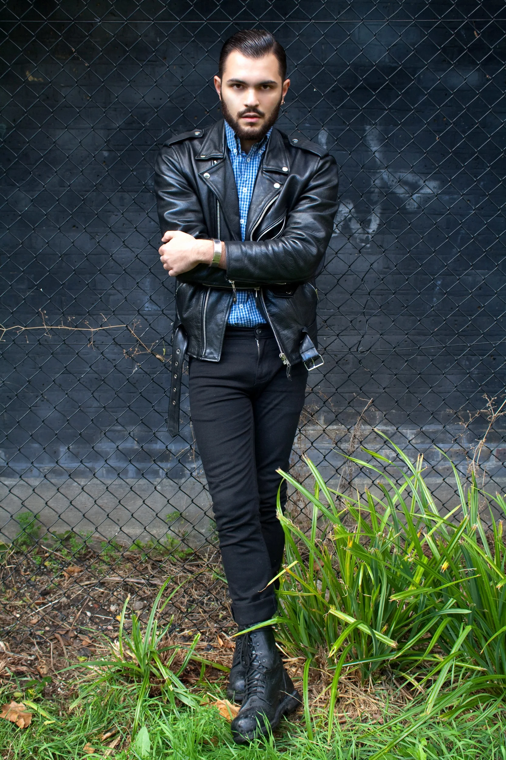Joven con ropa de cuero negro y botas, cruzado de brazos, posando frente a una cerca metálica negra en un fondo urbano.