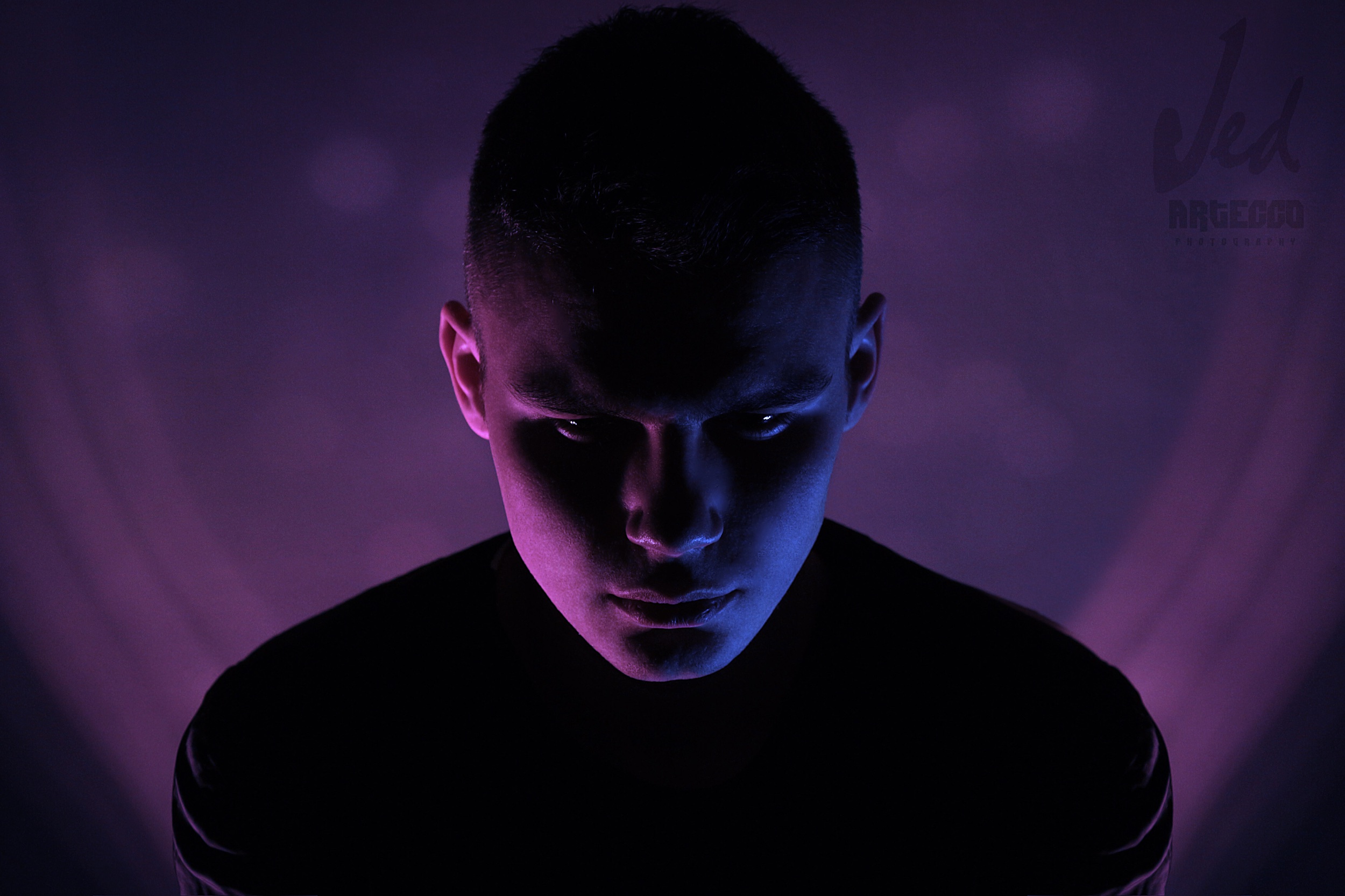 Retrato de un joven con iluminación morada y azul, con expresión seria y mirada hacia abajo, fondo oscuro.