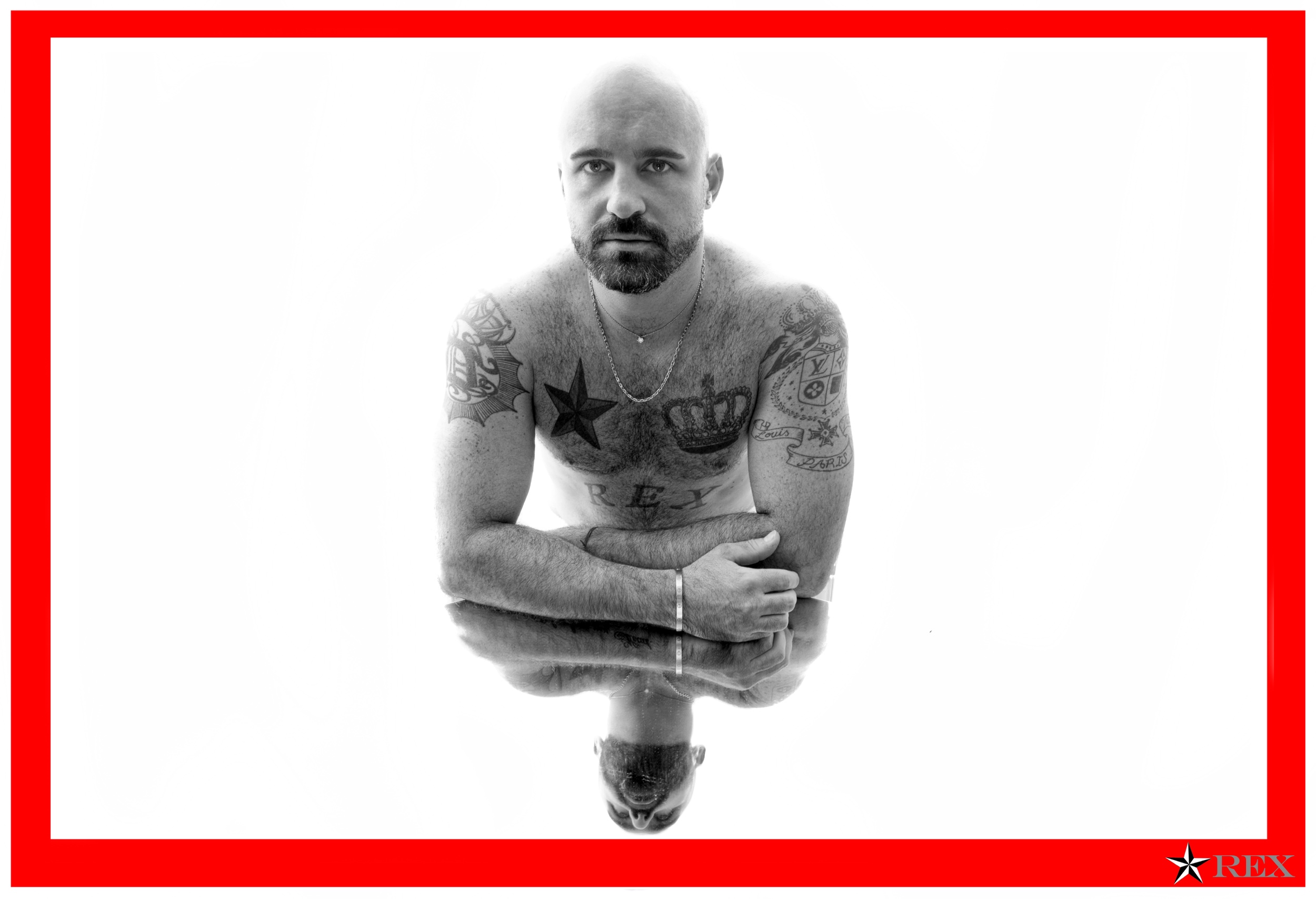 Hombre calvo sin camiseta con tatuajes en el pecho y brazos, con una cadena, en una pose inclinada con los brazos cruzados, reflejado en agua. Marco rojo con logo y texto 'REX' en la esquina inferior derecha.