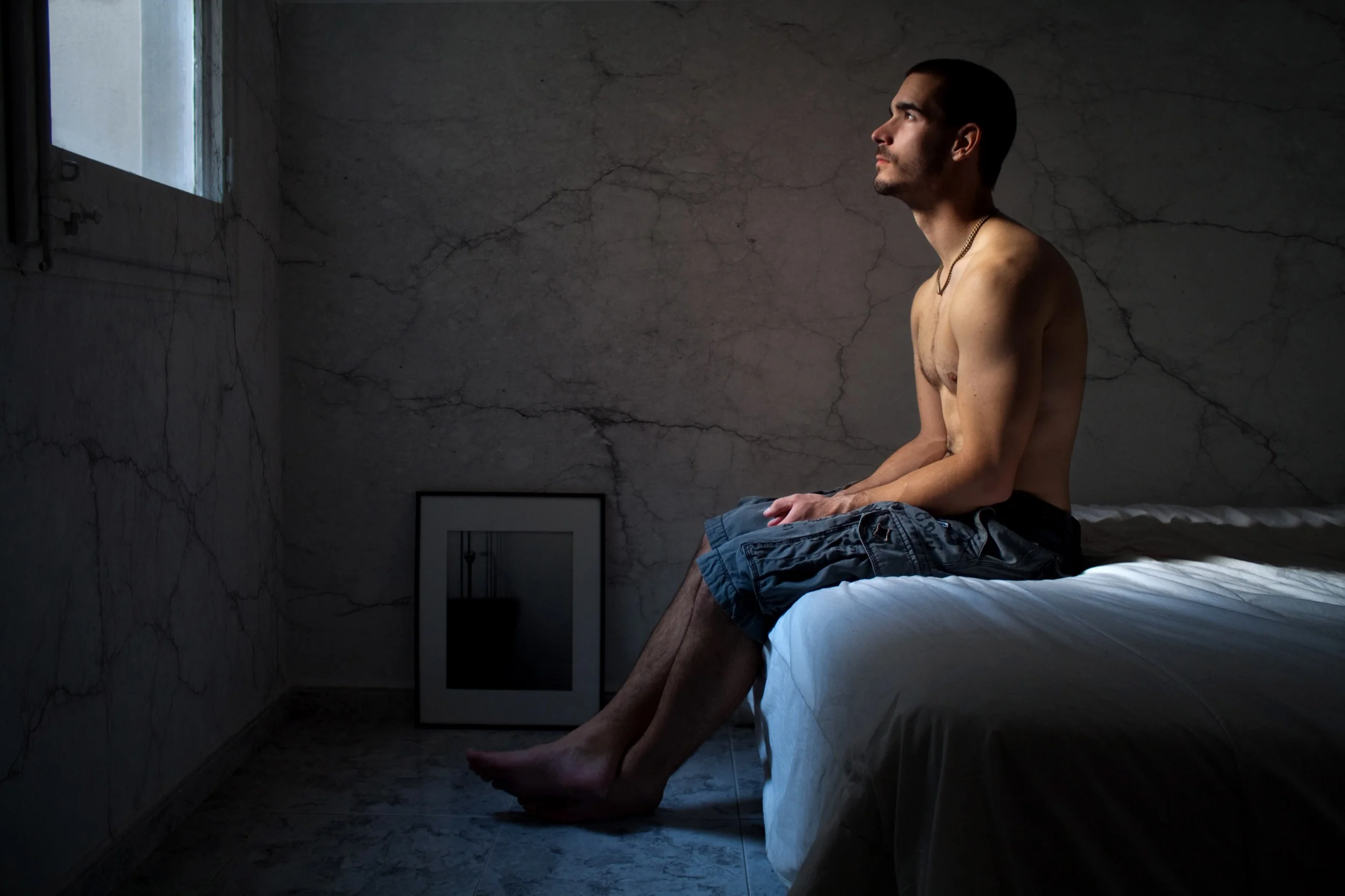Un hombre sin camiseta, sentado en un borde de la cama, mira por la ventana en una habitación con paredes de mármol y luz natural entrando por la ventana.