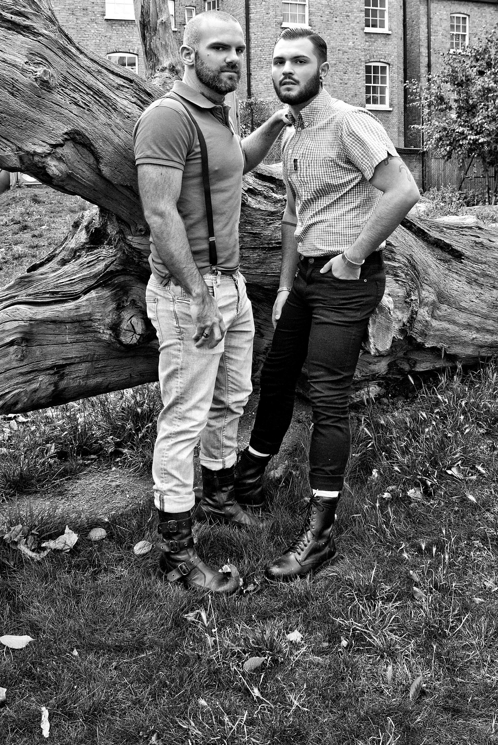 Dos hombres con barba y peinados estilosos, uno con pantalones claros y suspenders y el otro con pantalones oscuros, posando juntos en un jardín con árbol caído y edificios en el fondo, en una fotografía en blanco y negro.
