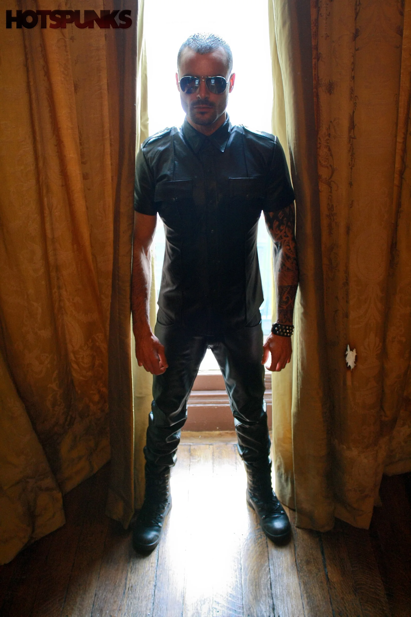 Hombre con gafas de sol, ropa de cuero negro, tatuajes en el brazo y botas, posando en un círculo de luz natural entre cortinas doradas.