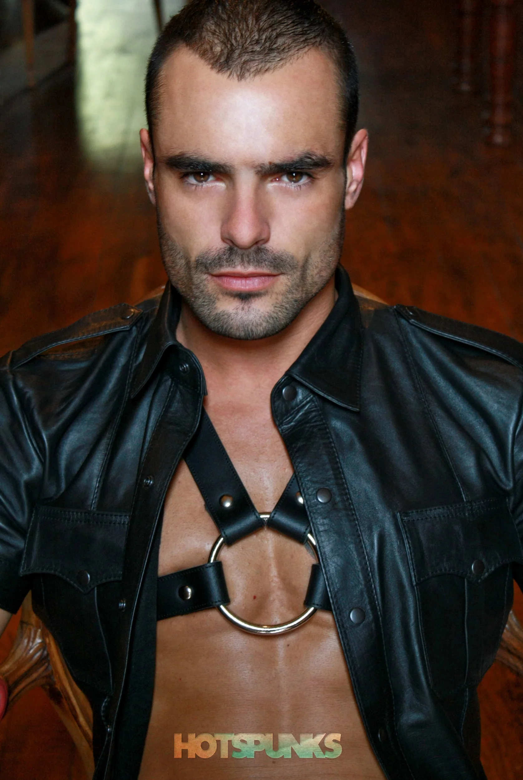 Hombre con barba, sin camisa, con un arnés de metal en el pecho, lleva chaqueta de cuero negro, en un entorno interior.