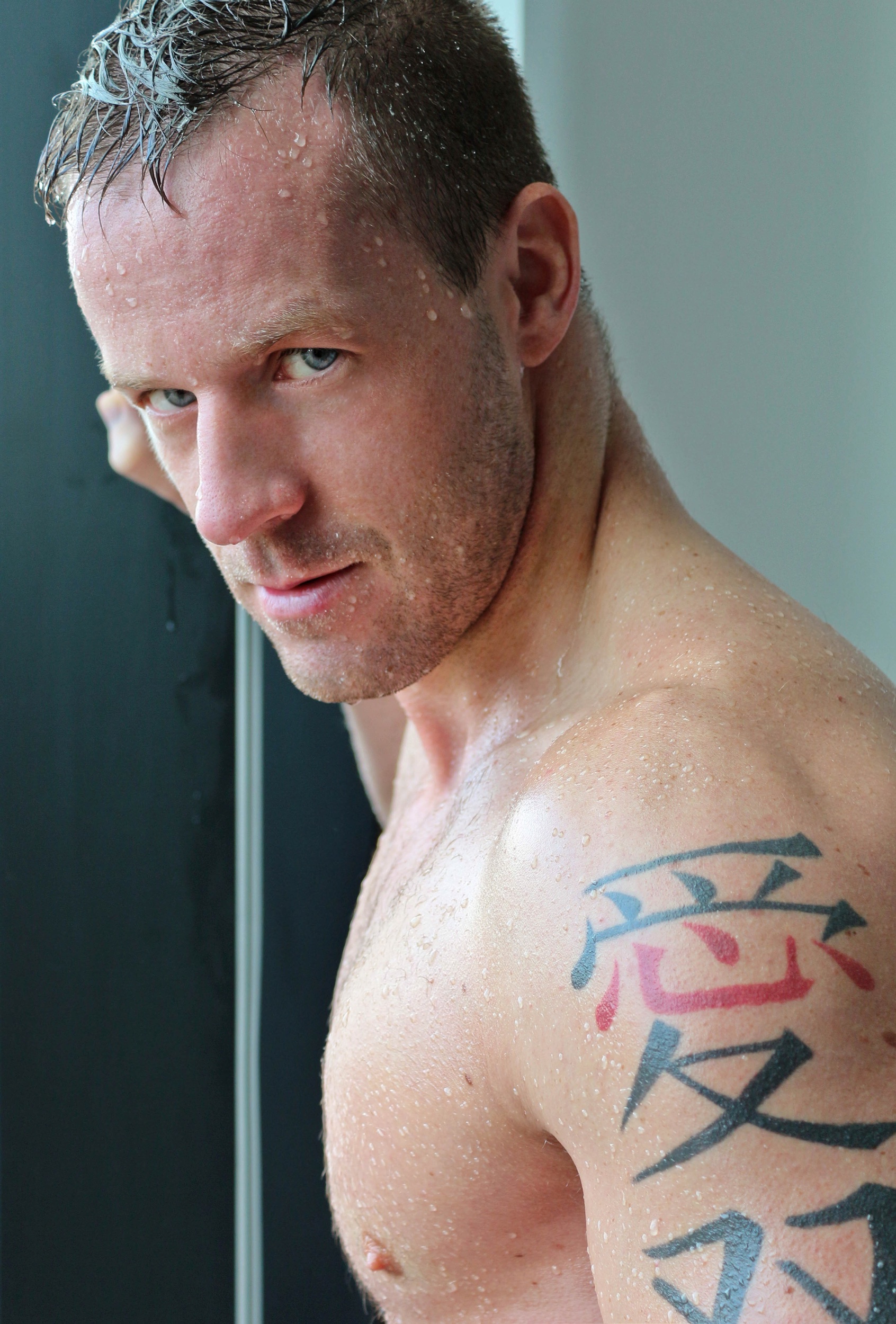 Hombre con cabello húmedo, piel mojada y tatuaje en el hombro, mirando hacia la cámara