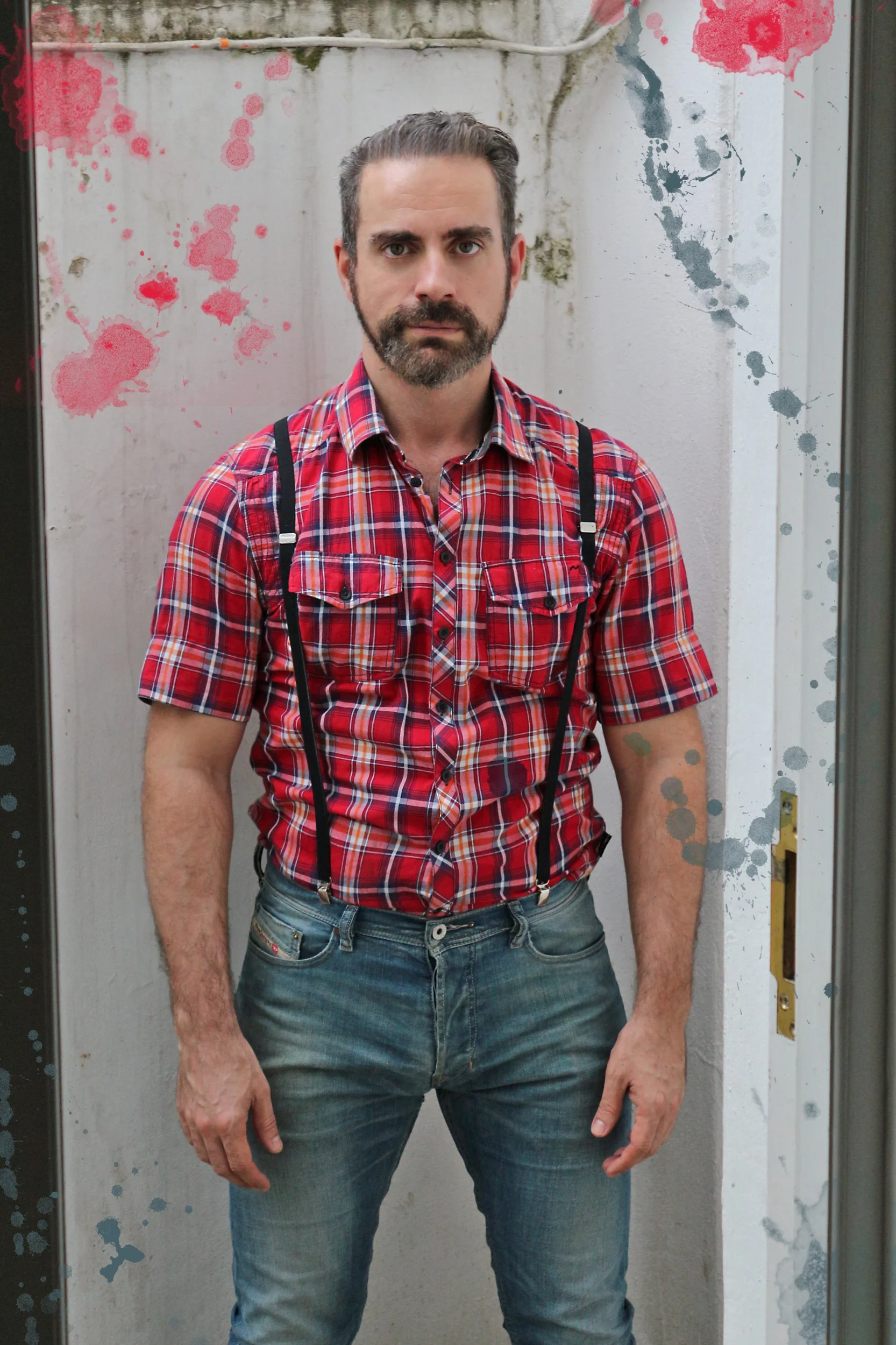 Hombre con barba y cabello peinado, usando camisa de cuadros rojos y pantalones vaqueros, de pie en un ascensor con manchas de pintura en las paredes.