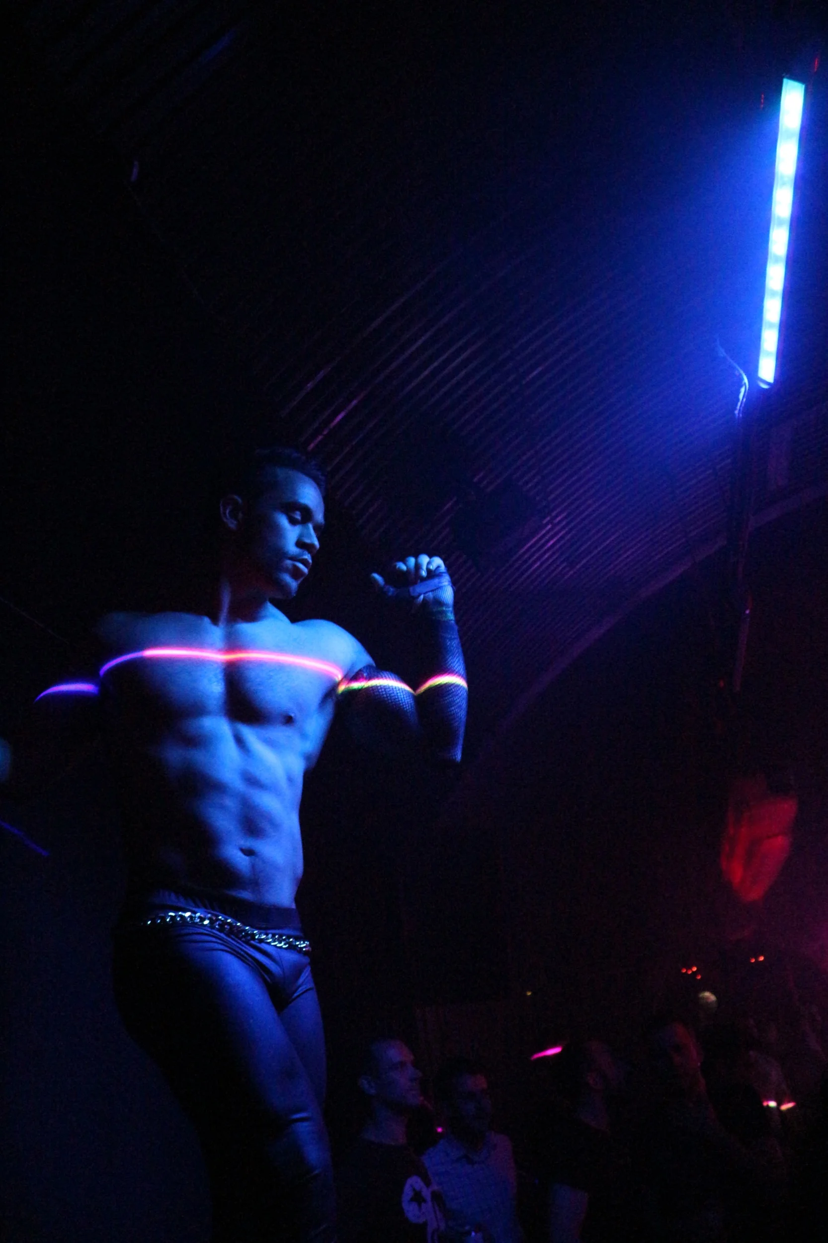 Un hombre con músculos visibles, vestido con pantalones ajustados y con una cadena, brilla bajo luces UV en un ambiente oscuro, posiblemente en un club nocturno, rodeado de personas.