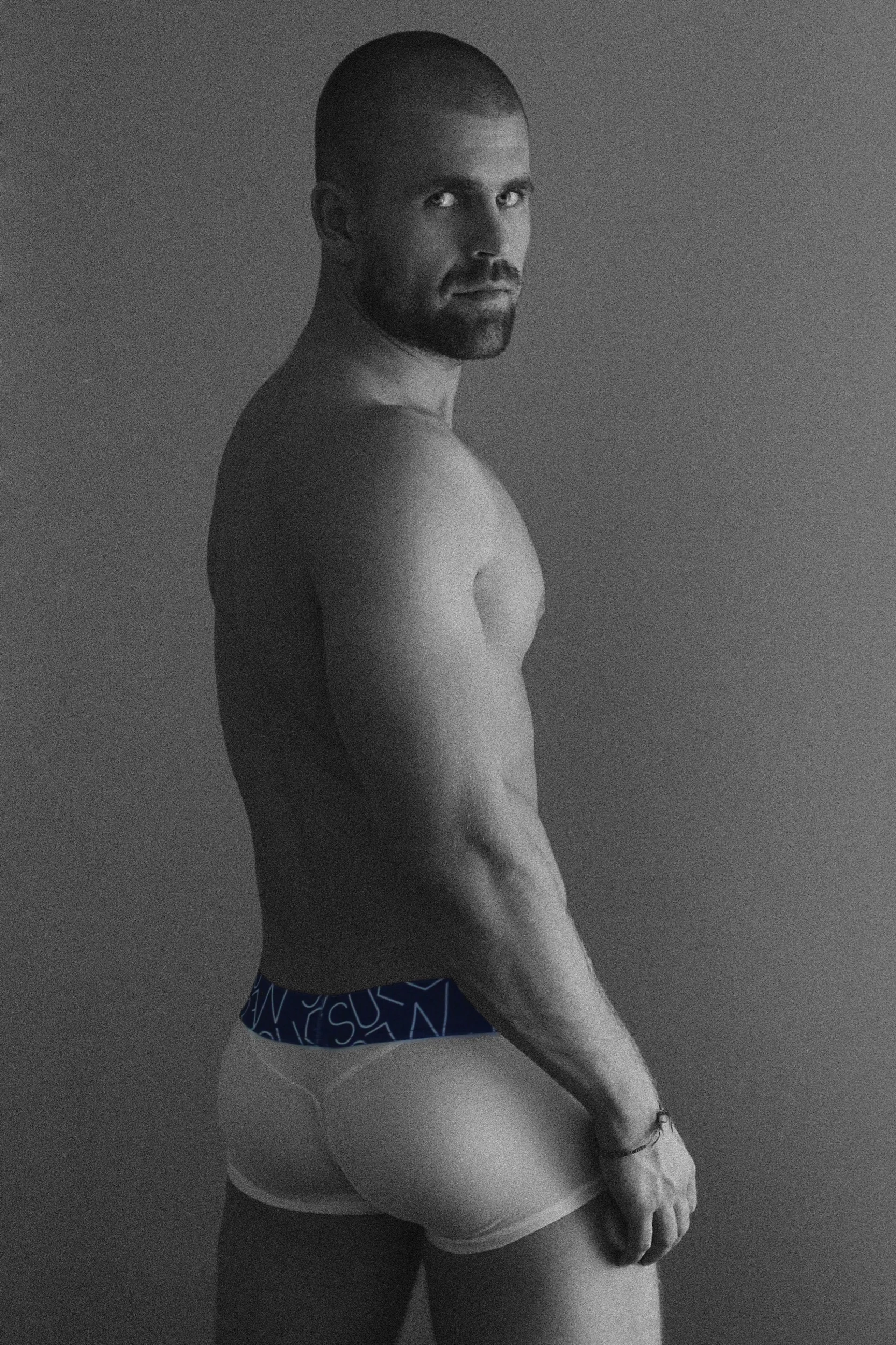 Hombre desnudo de espaldas con boxer azul y ceñido, mirando hacia la cámara, en fondo gris.