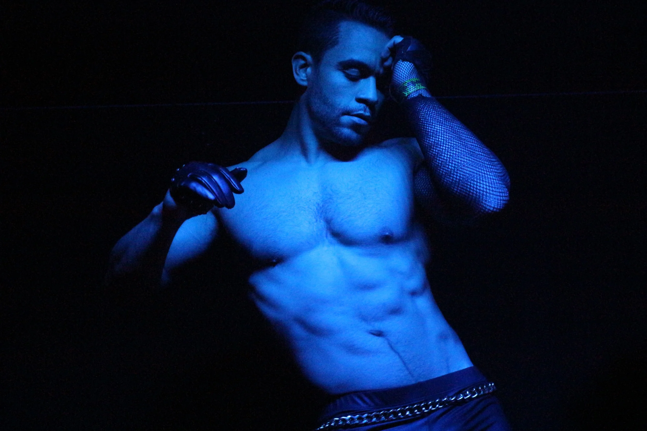 Hombre musculoso desnudo bajo iluminación azul, con guantes de látex y manga de red en un fondo oscuro.