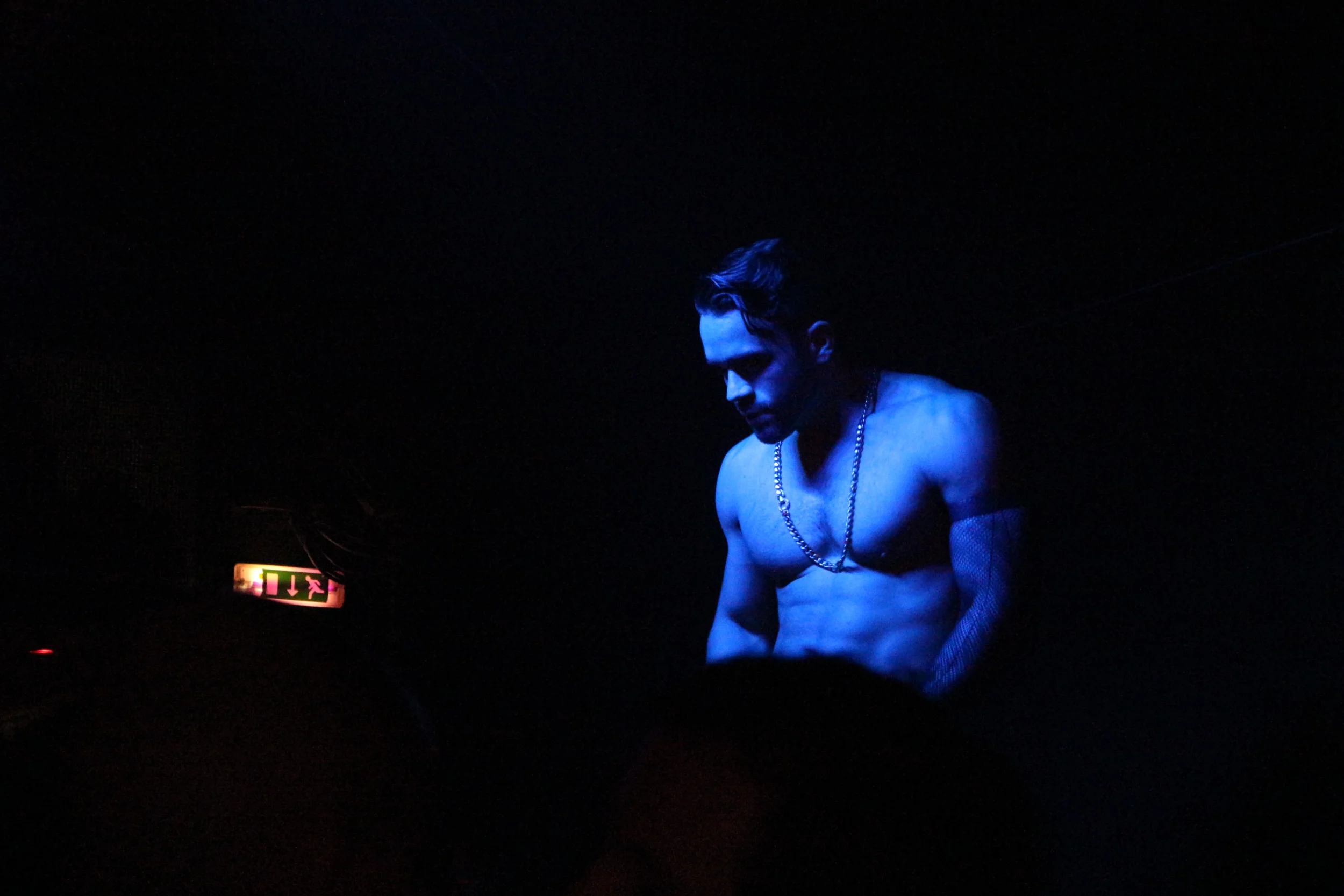 Un hombre sin camisa con tatuajes, usando cadenas, en un ambiente oscuro y con iluminación azul, probablemente en un evento o espectáculo.