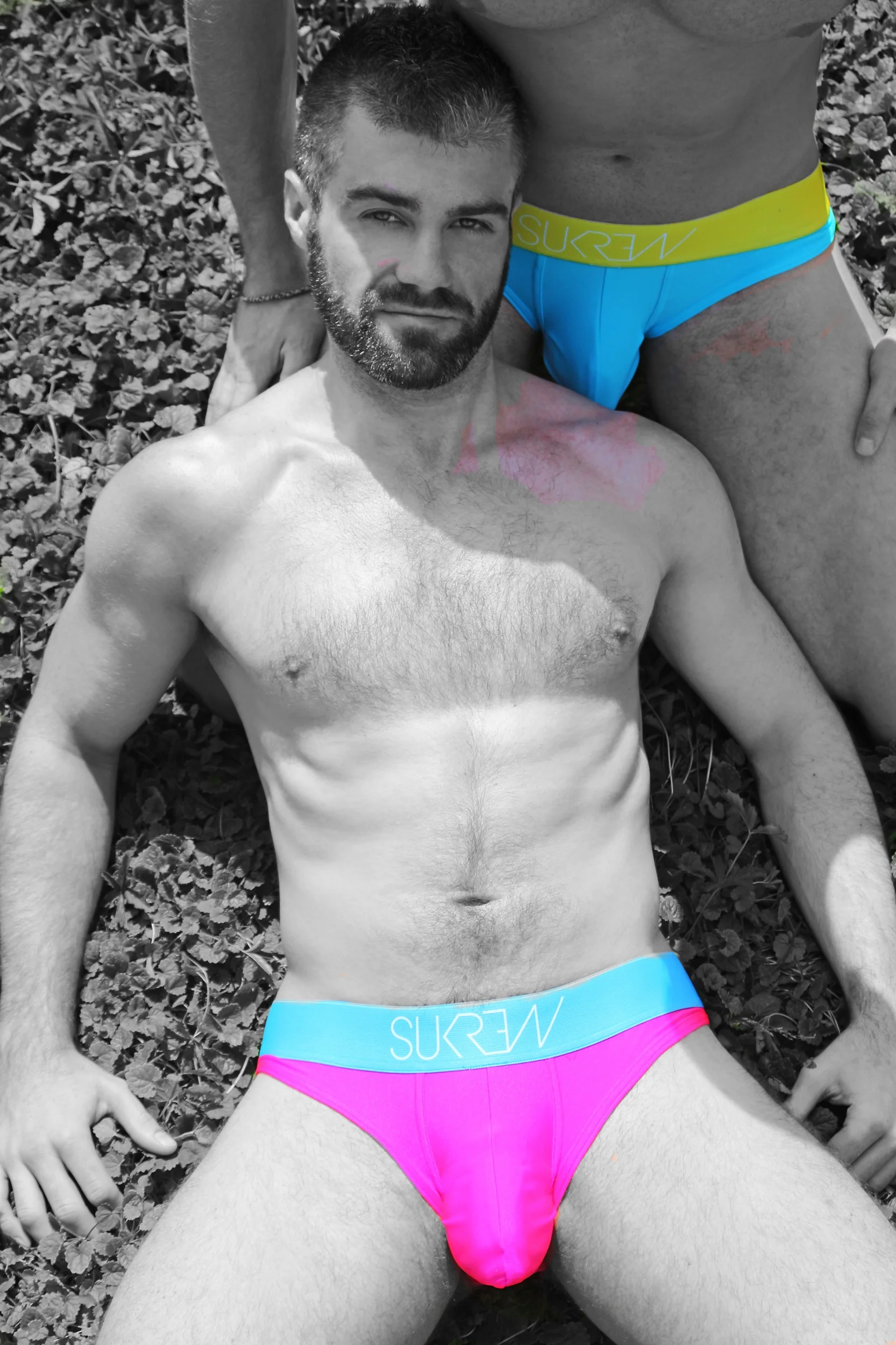 Dos hombres en ropa interior, uno está acostado en la hierba y es en su mayoría en color negro y blanco, mientras que el otro está sentado y lleva ropa interior azul y rosa. La imagen tiene un filtro en escala de grises con los colores del otro hombr