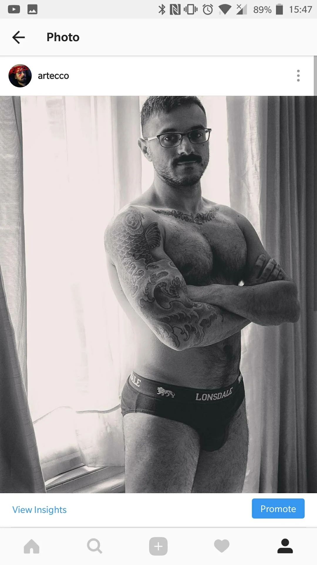 Hombre con barba y gafas de pie con brazos cruzados en ropa interior, mostrando tatuajes en brazos y torso, frente a cortinas de fondo en una habitación