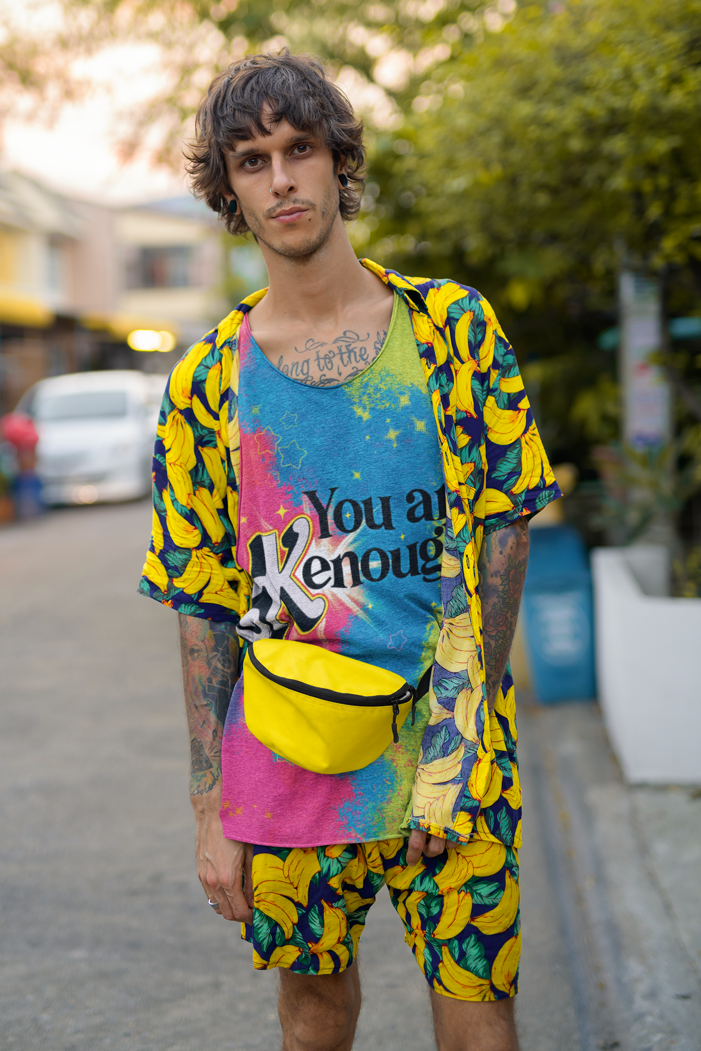 Joven con peinado desordenado, ropa colorida con estampados de plátanos, usando una camiseta que dice "You are Enough", con una riñonera amarilla, en una calle con árboles y autos en el fondo.