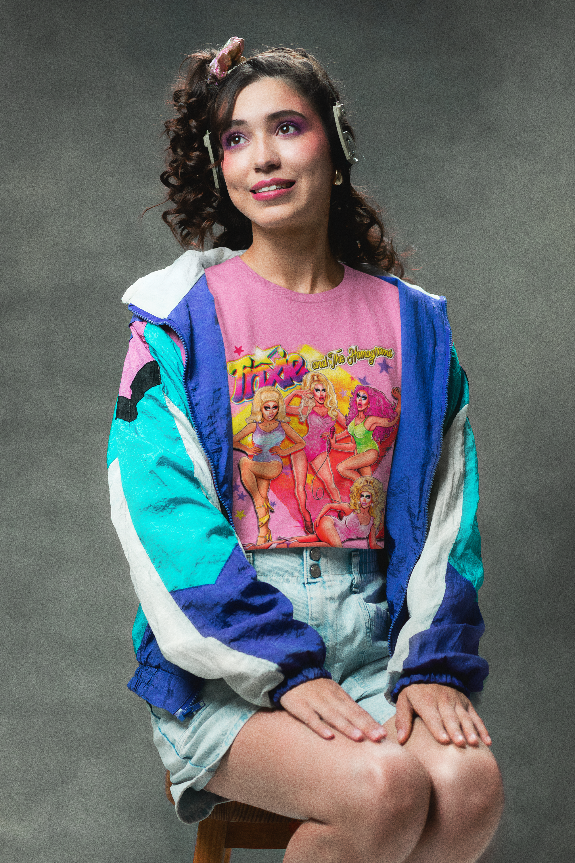 Joven mujer con cabello rizado y maquillaje colorido, usando un overol y camiseta con ilustraciones de las Toyz y las Flameammers, sentado en un banco, en un fondo gris.