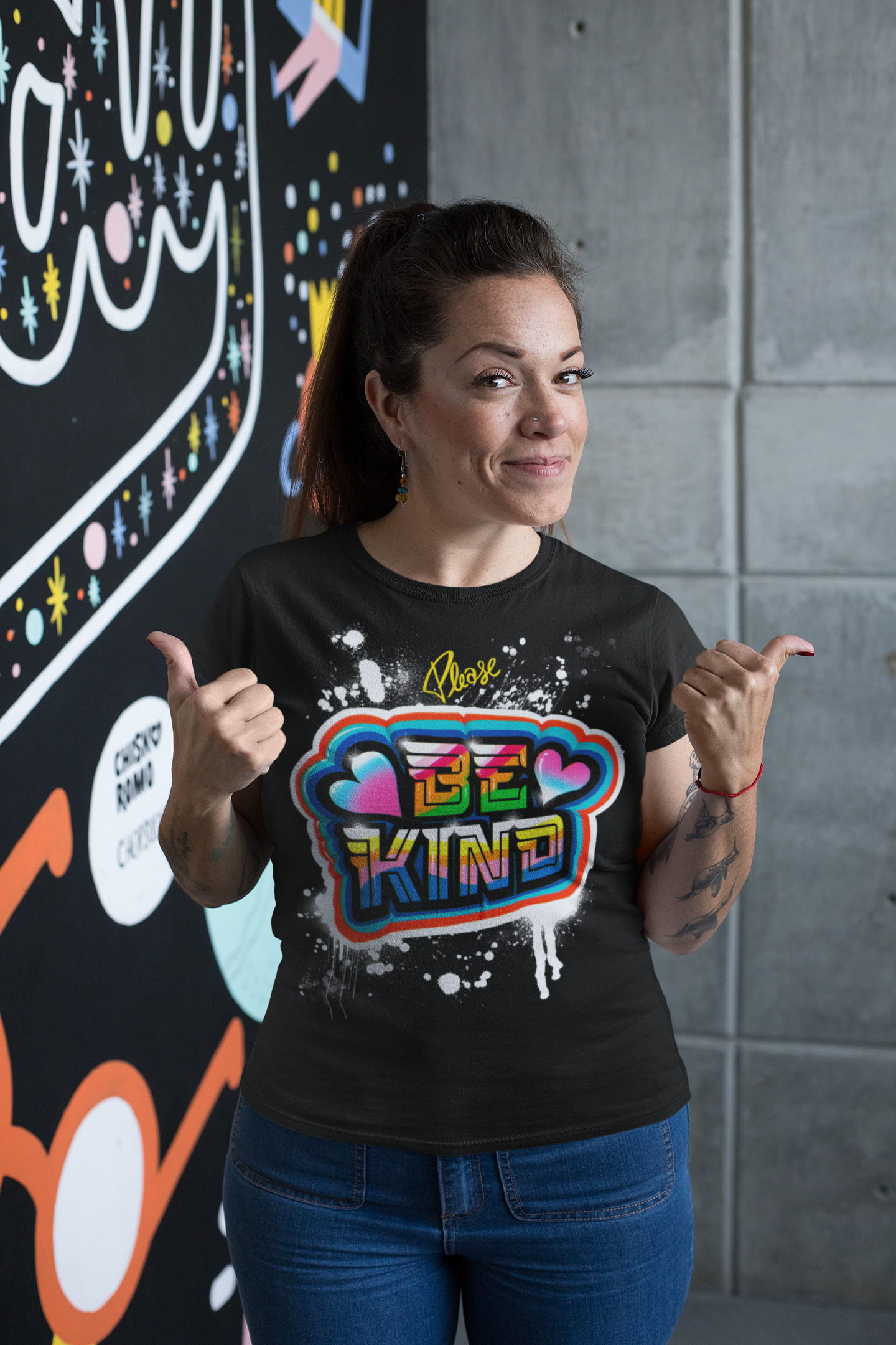 Mujer sonriendo y haciendo señal de aprobación con ambas manos, usando una camiseta con el texto 'Please Be Kind' y decoraciones coloridas, en un fondo con arte mural.