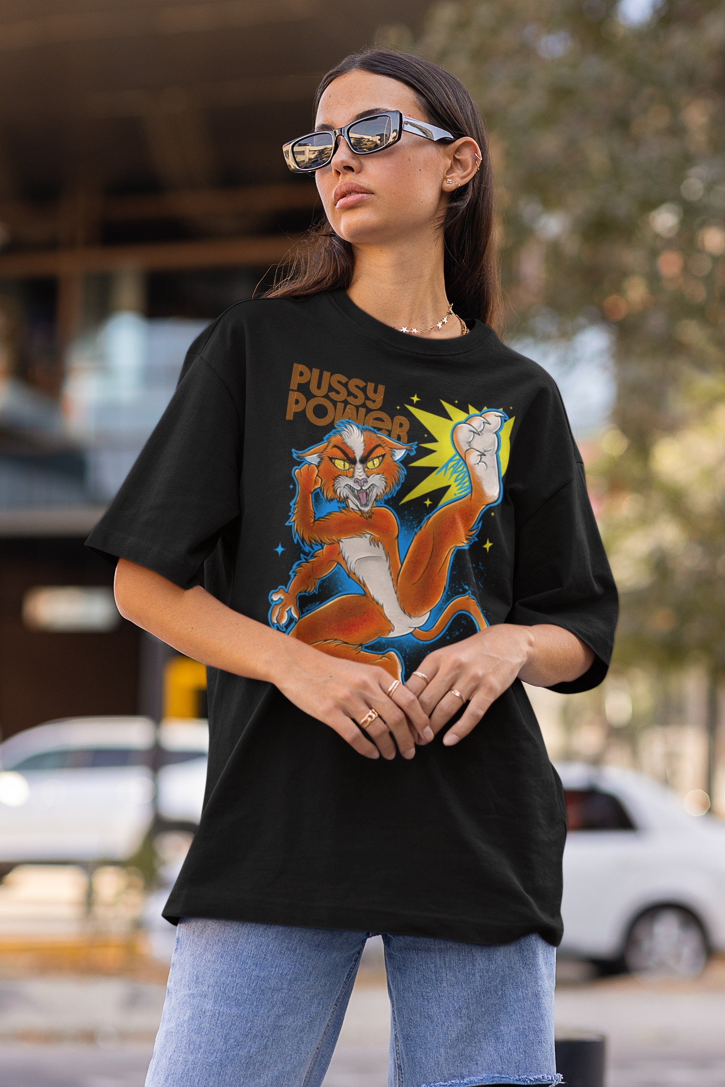 Mujer con gafas de sol oscuras y camiseta negra con un diseño de un león caricaturesco y texto 'Pussy Power', en un entorno urbano con árboles y autos de fondo.