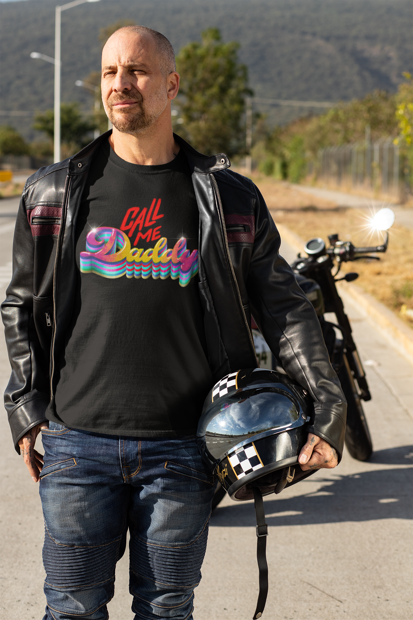 Hombre con chaqueta de cuero, camiseta con el texto 'Call Me Daddy', sosteniendo casco de motocicleta, de fondo una motocicleta y árboles, en un día soleado.