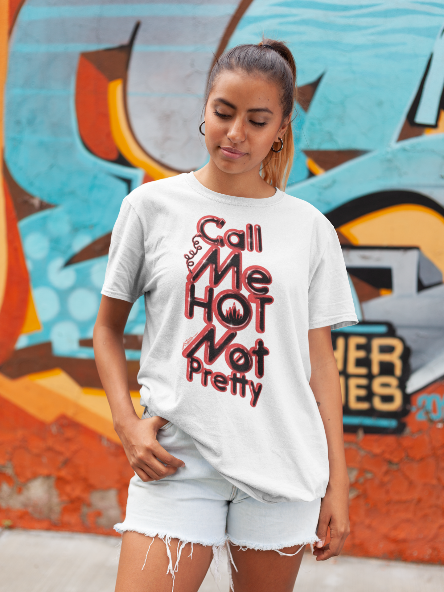 Joven mujer con camiseta blanca que dice 'Call me hot, not pretty' en un mural de graffiti colorido.
