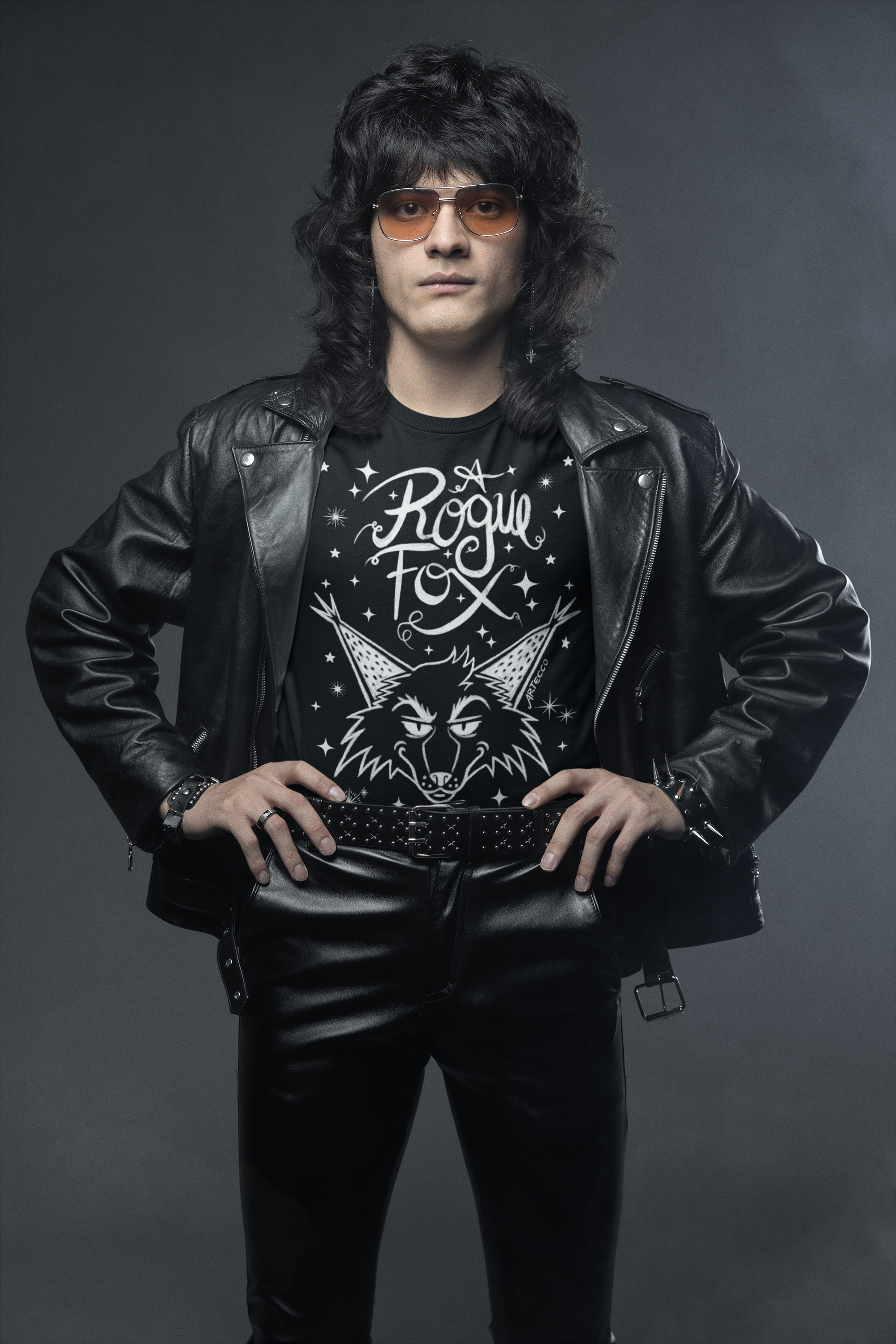 Joven con cabello largo y rizado, gafas de sol, chaqueta de cuero negra, camiseta con diseño de un lobo y la frase 'A Rogue Fox', y pantalones de cuero, posando con las manos en las caderas contra un fondo gris oscuro.