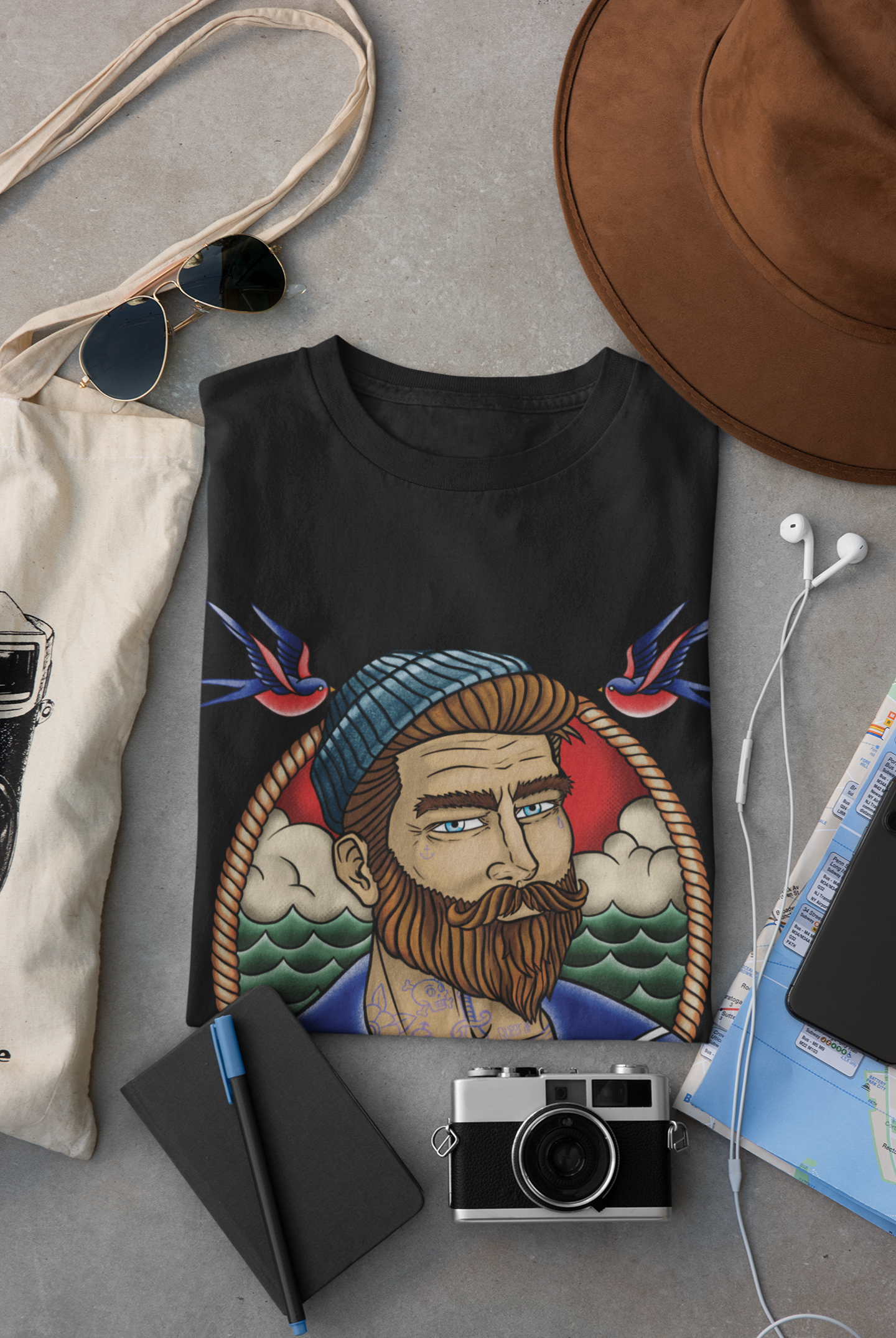 Camisa negra con diseño de un hombre con barba y tatuajes, con gorro y lágrimas, rodeado de nubes y olas, flanqueado por dos colibríes. Rodeado de objetos de viaje como sombrero, gafas de sol, mochila, cámara, libro, mapa, auriculares y teléfono.