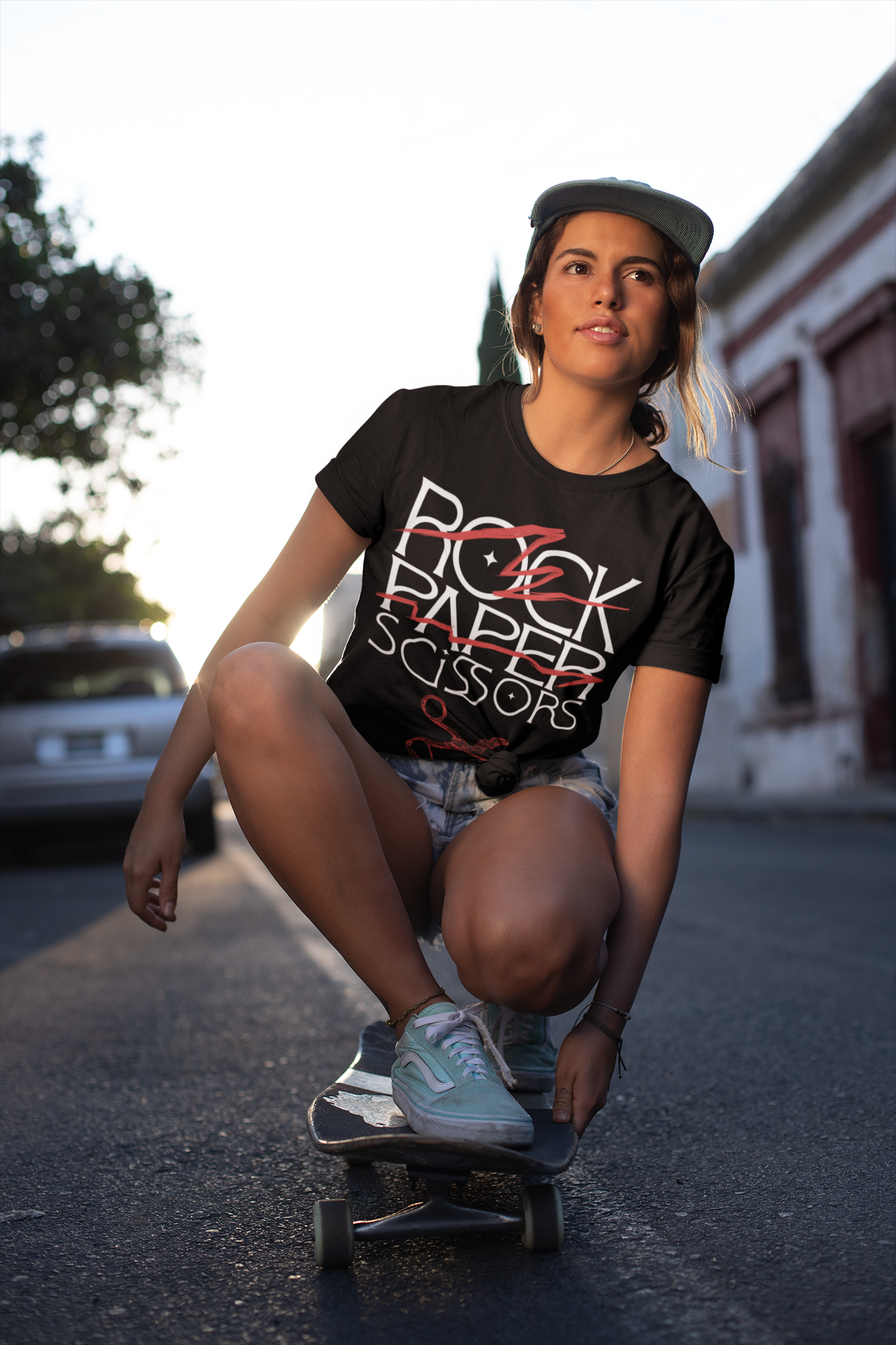 Joven mujer montando en patineta en la calle durante el atardecer, vestida con ropa casual y gorra, con edificios antiguos y árbol en el fondo.
