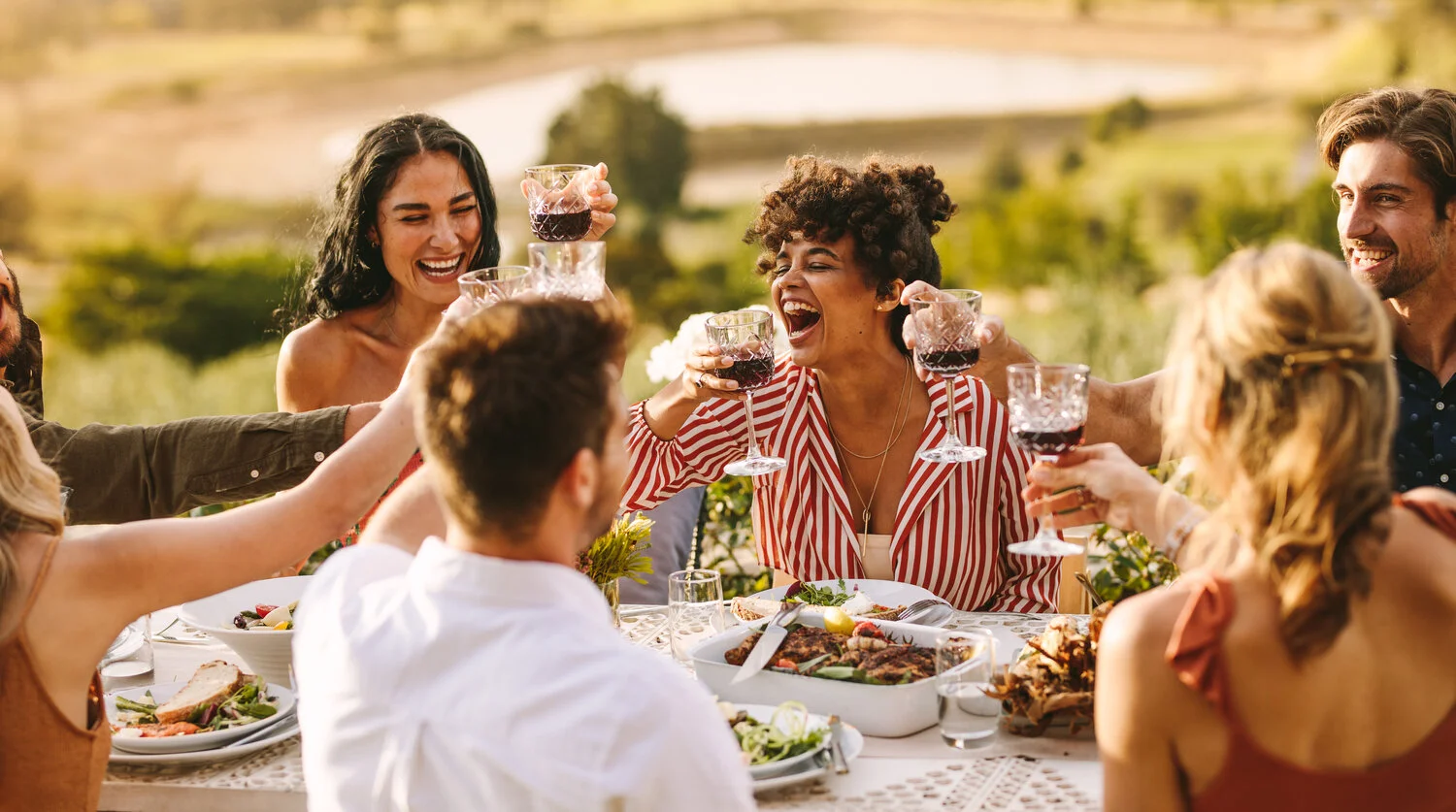 A group of young friends laughing as they toast with red wine over a table full of food