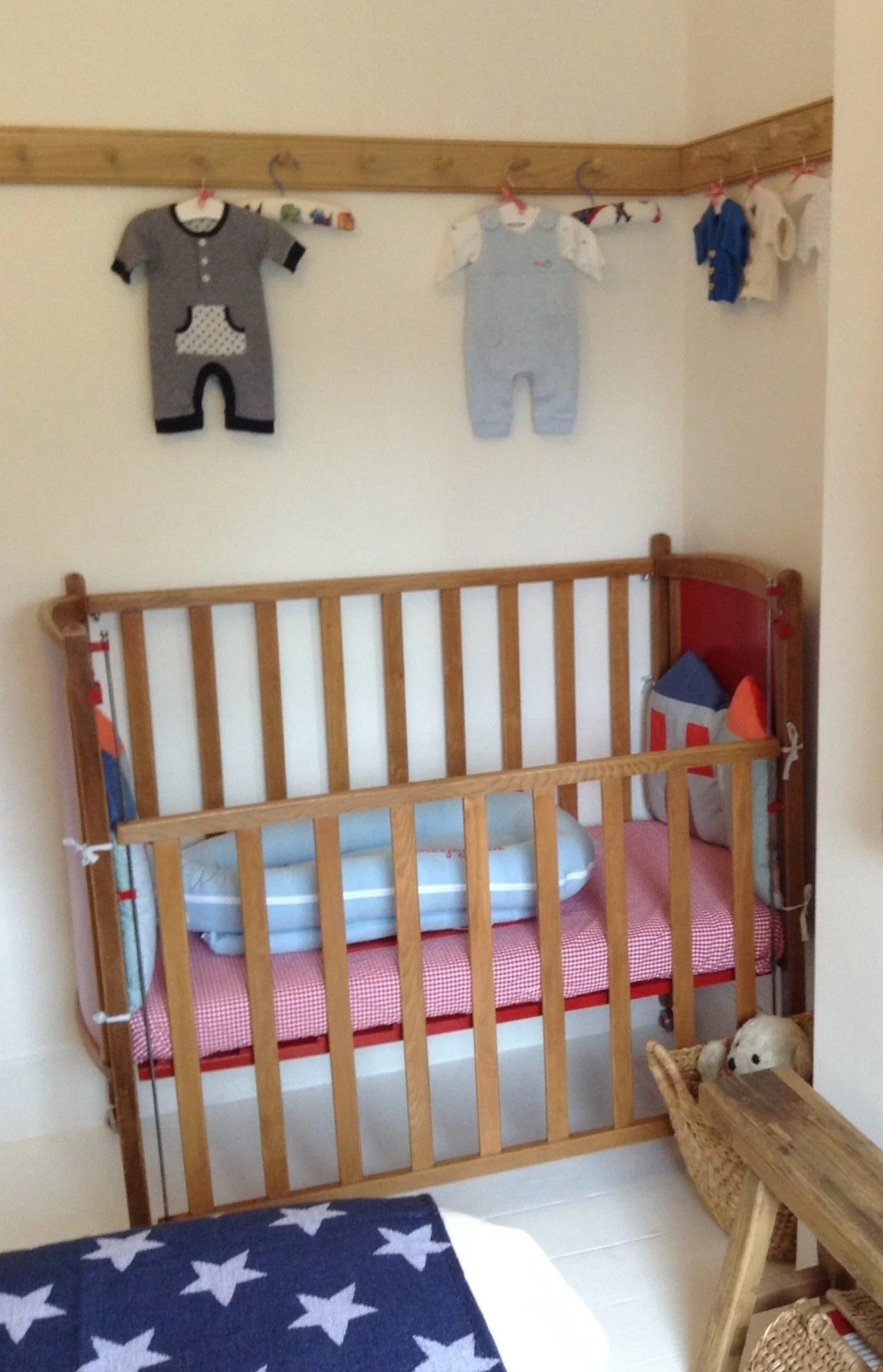 Cot painted red along a white wall underneath a peg rail with baby clothes hanging