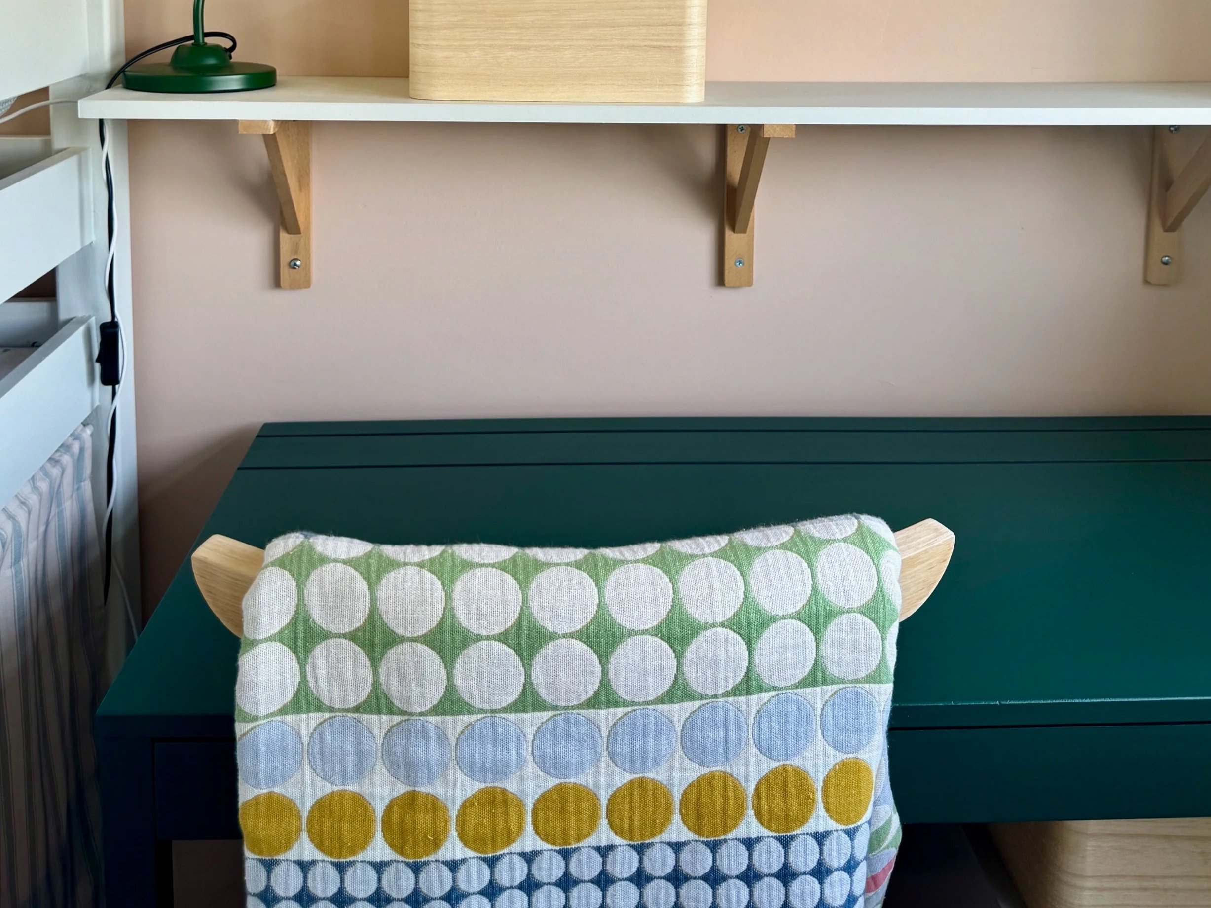A green table with a patterned cloth featuring large circular dots in green, yellow, blue, and white. Above the table, a wooden shelf with a green desk lamp on the left, a light wooden box in the middle, and an empty space on the right.