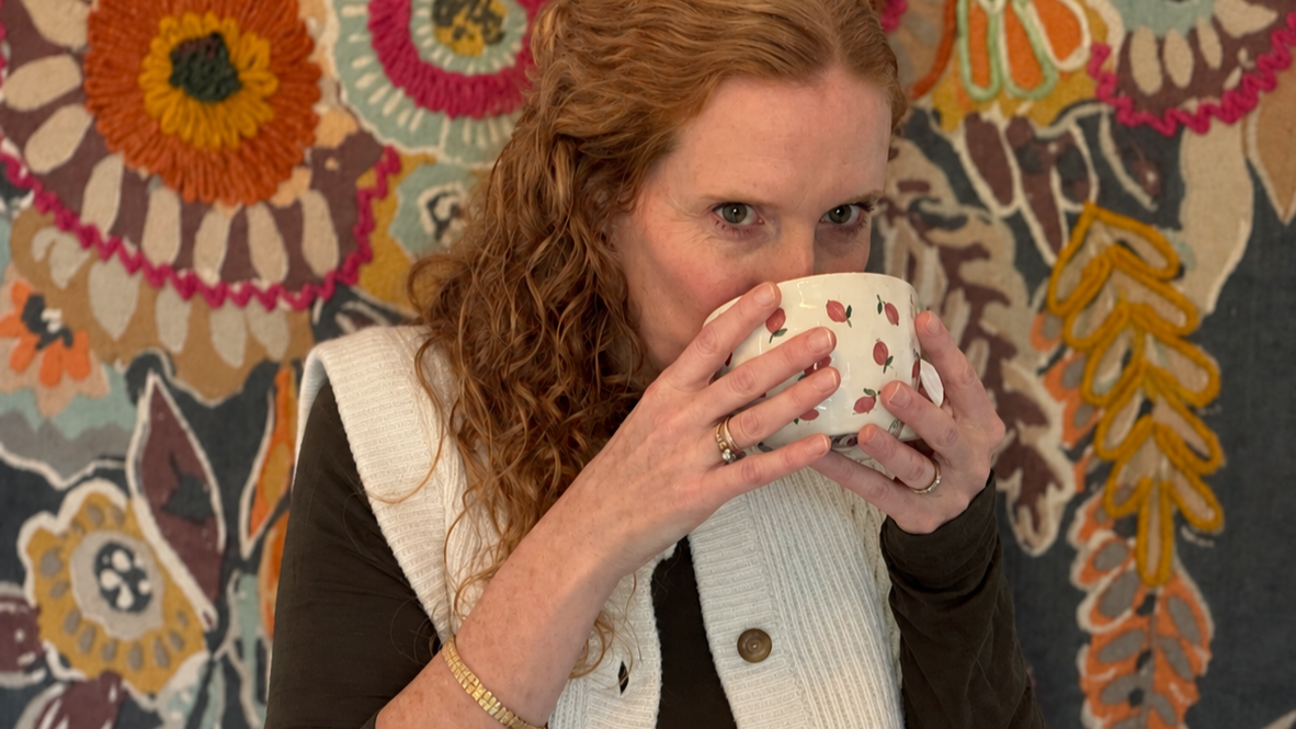 A woman with red curly hair sipping from a white mug decorated with pink cherries, standing in front of a colorful, floral-textured tapestry.