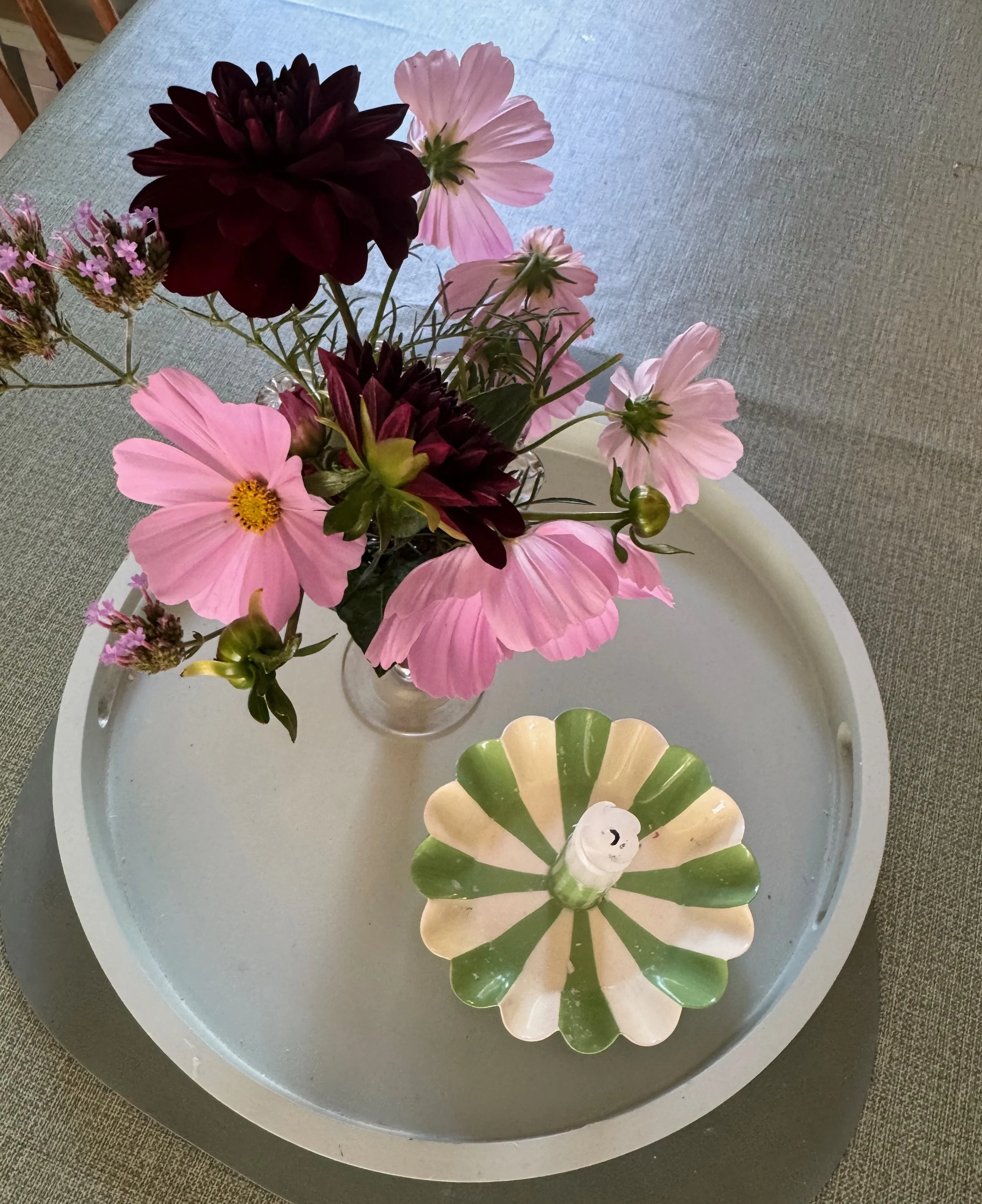 A floral arrangement in a small glass vase on a light gray table. The flowers include large pink blossoms, dark burgundy flowers, smaller pink flowers, and a small branch with purple flowers.