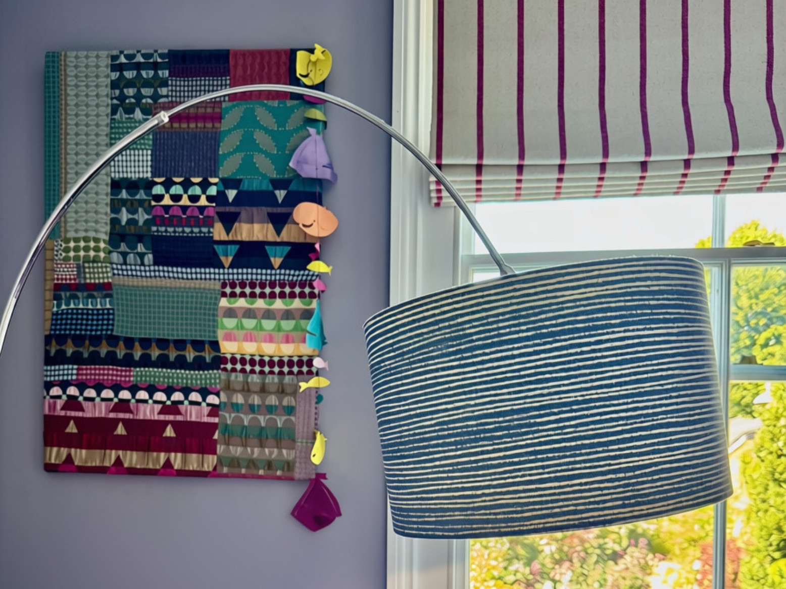 Interior view of a room with a colorful fabric art piece on the wall, a window with a striped Roman shade, and a modern lamp with a curved arm and a blue and white striped lampshade.