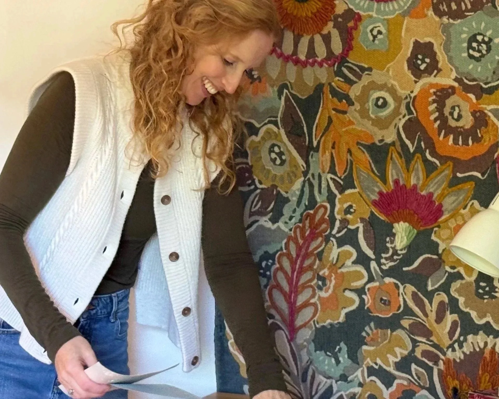 A woman with curly red hair wearing a cream vest over a black top and blue jeans, smiling while looking down at her left hand, standing beside a large, colorful floral-patterned textile wall art.