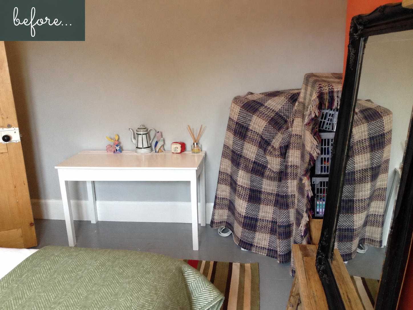 Room with. grey floor, small white desk and a pile of storage boxes hidden under a rug