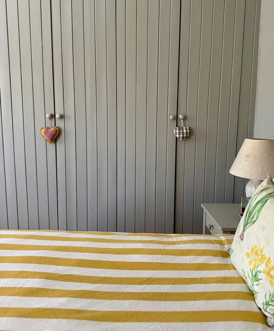 A bedroom with a bed covered in a yellow and white striped bedspread, a pillow with a floral print, a nightstand with a lamp, and a closet with two doors decorated with heart-shaped ornaments.
