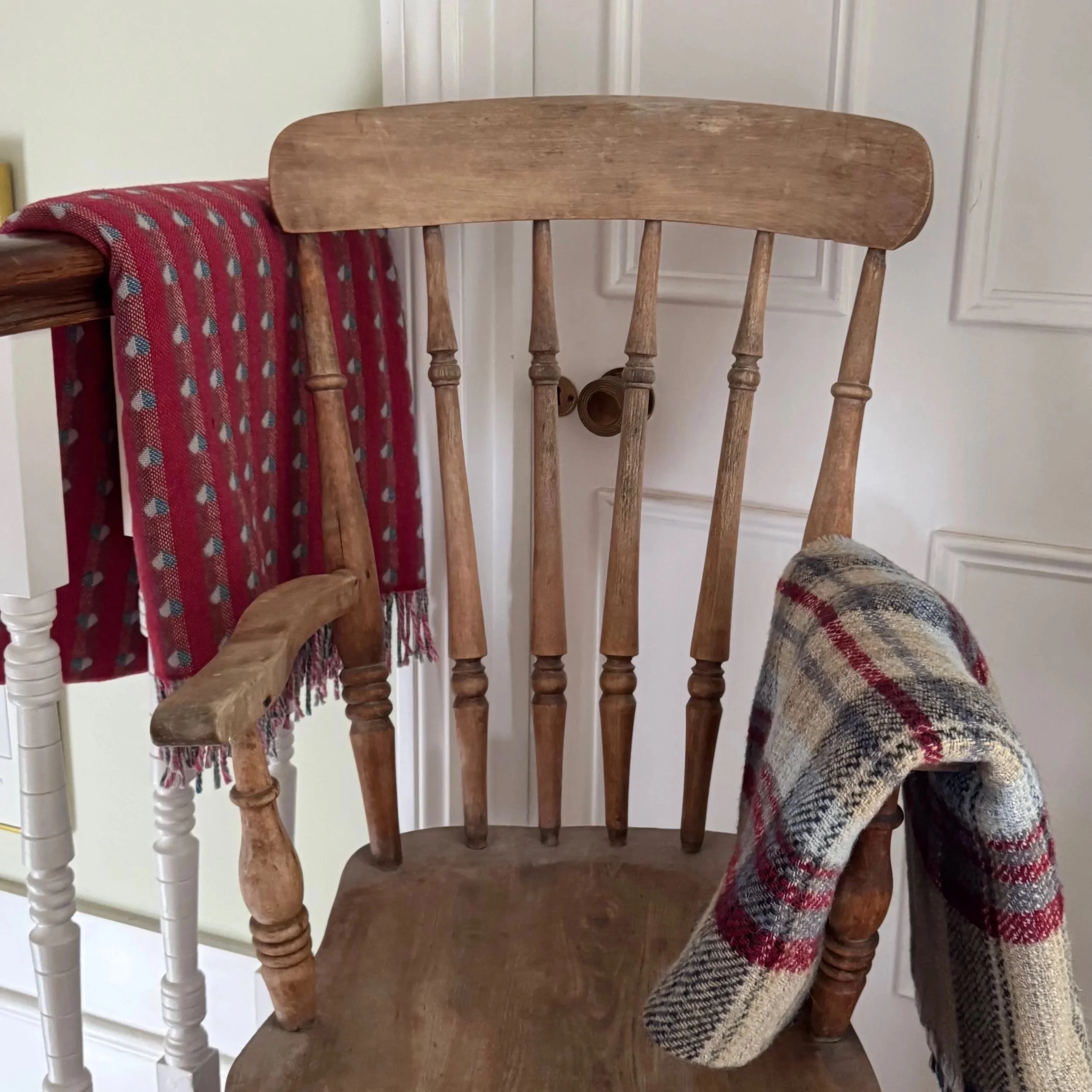 An empty wooden dining chair with turned legs and a curved backrest, placed next to a table with a red and blue plaid blanket hanging over the side, in a room with white paneled walls.