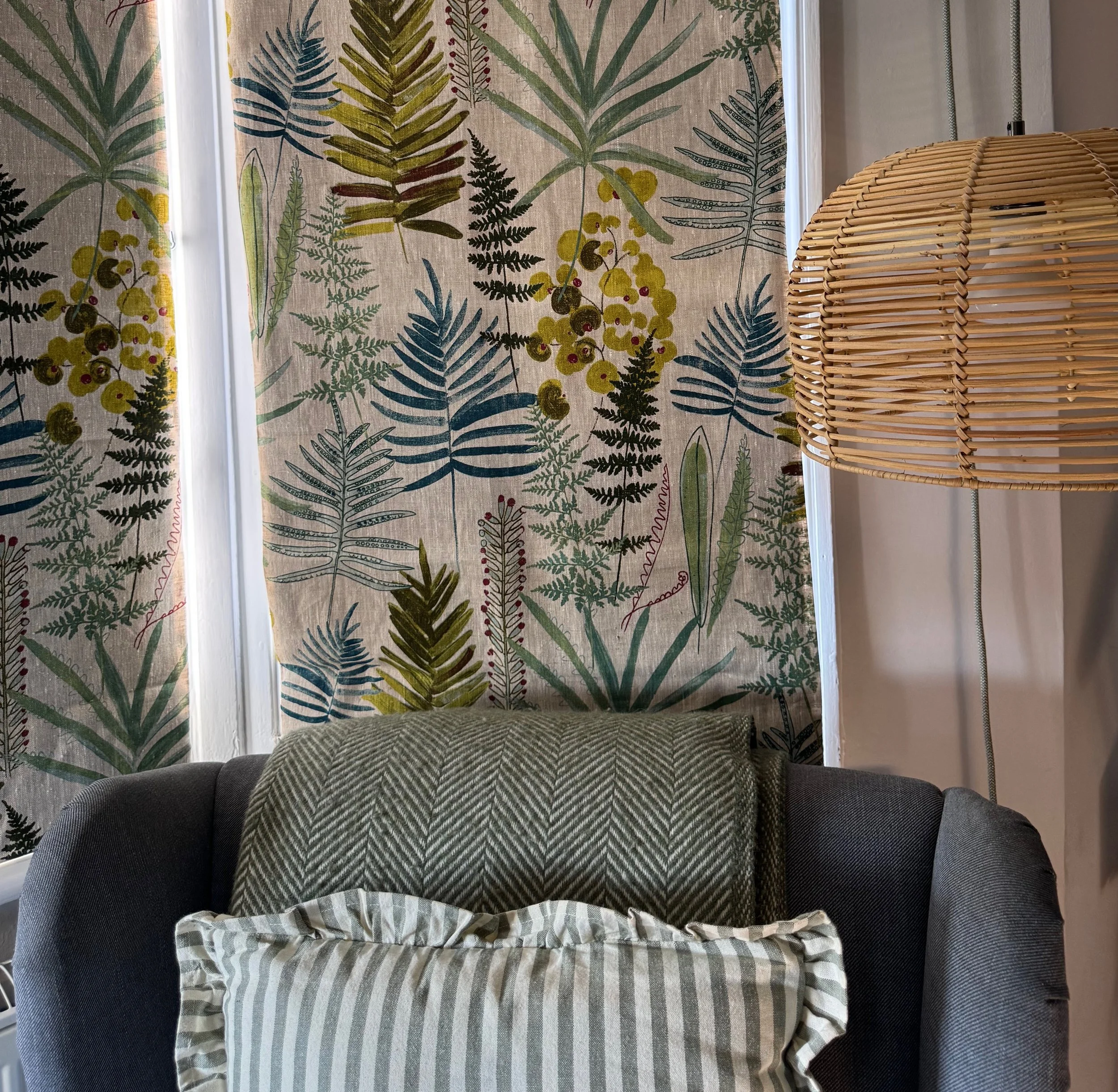 View of curtains with a botanical pattern overlooking a window, a wicker lampshade, and a couch with a striped blanket in a living room.