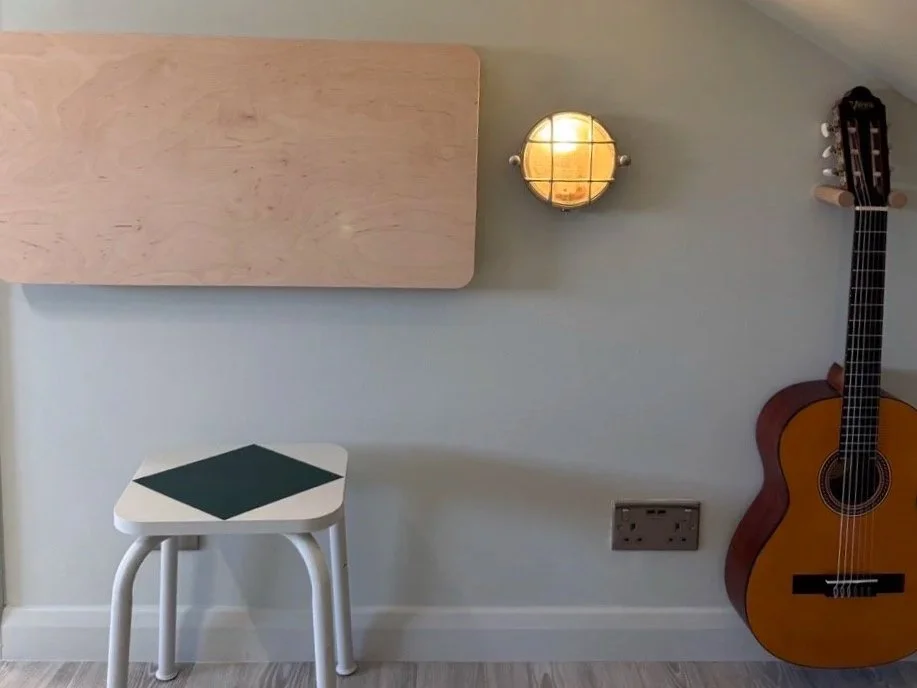 A cozy corner with a small stool, a wooden guitar, a wall light, a wall-mounted wooden folding desk