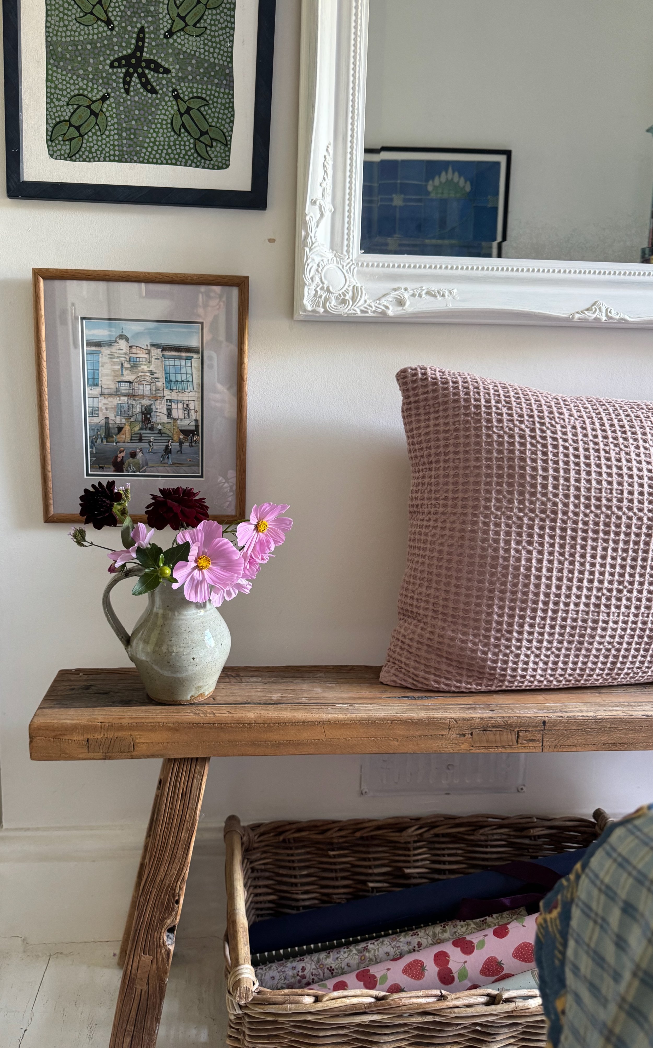 A wooden table with a beige ceramic pitcher holding pink and dark red flowers, a pink textured pillow on a wall, framed pictures on the wall, a wicker basket with a strawberry-patterned cushion, and a mirror with an ornate white frame.