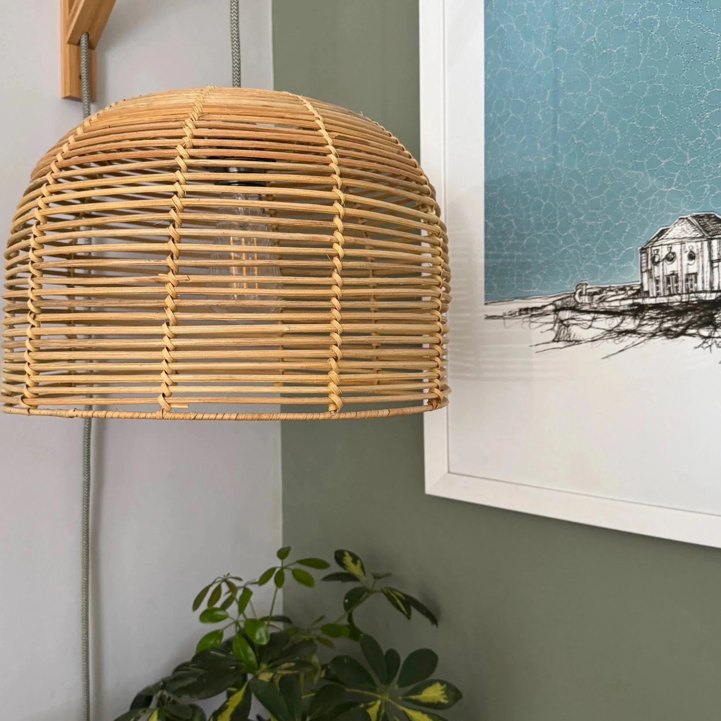 Close-up of a woven rattan lamp with a plant in the background against a gray wall and a framed picture.