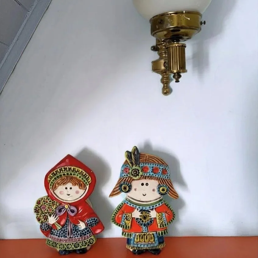Two colorful folk art dolls, one dressed in a red hooded cloak and the other in traditional costume with a headpiece, placed on a surface with a brass wall-mounted fixture above them.