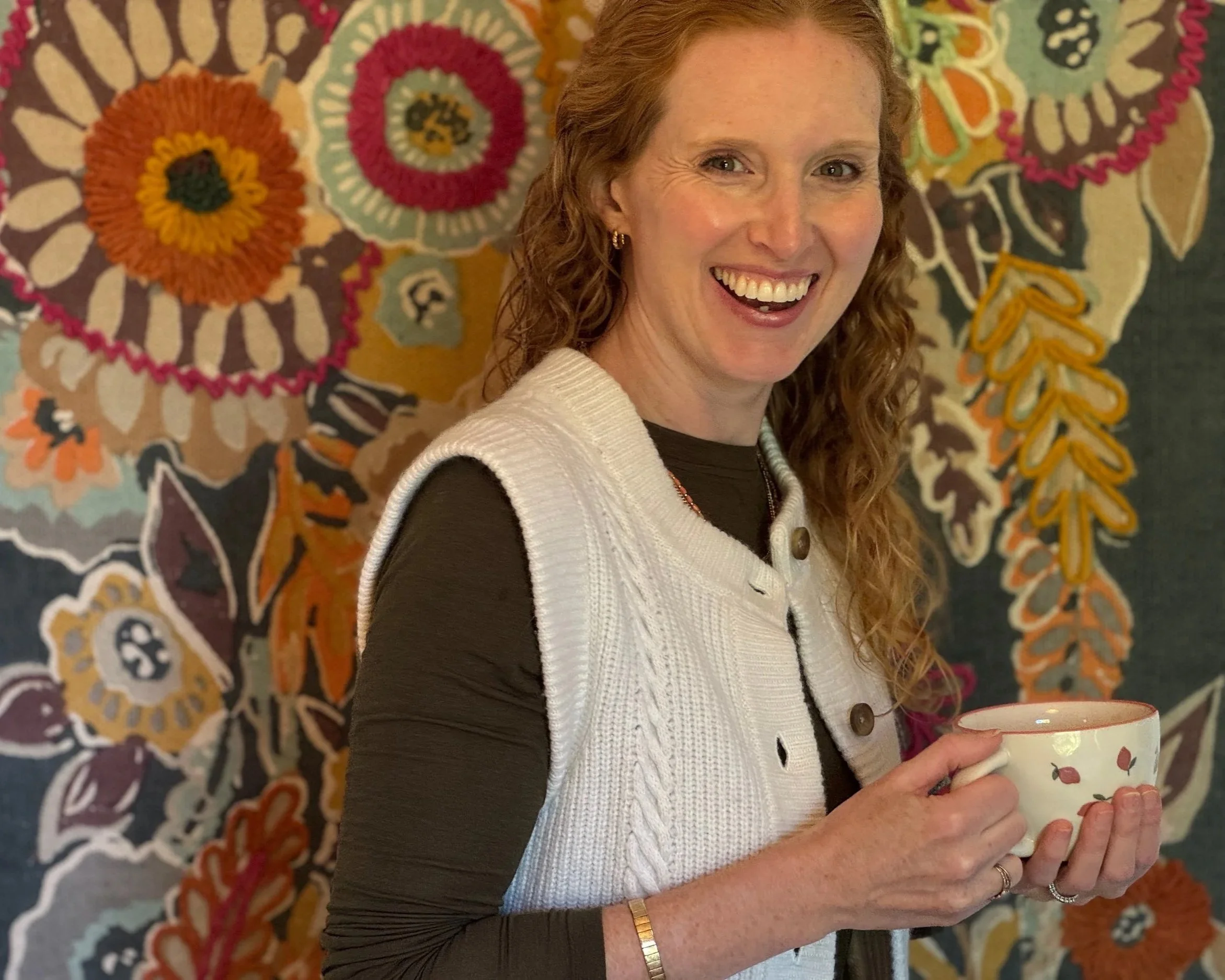A woman smiling while holding a floral patterned mug, standing in front of a colorful, intricate tapestry background.