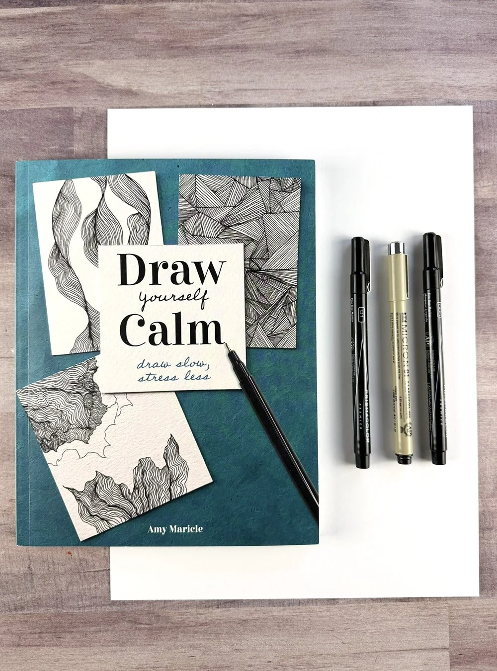 A copy of the book, "Draw Yourself Calm" by Amy Maricle with 3 pens on a desktop with some white paper.