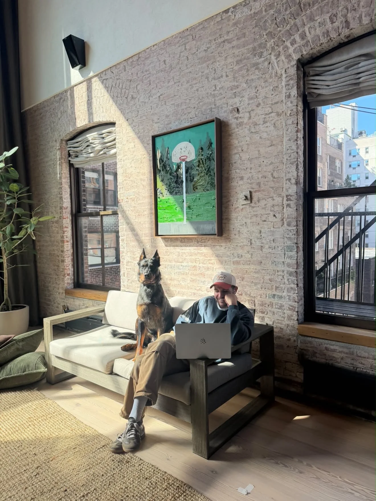 A man works on a laptop beside a Beauceron resting on the couch.
