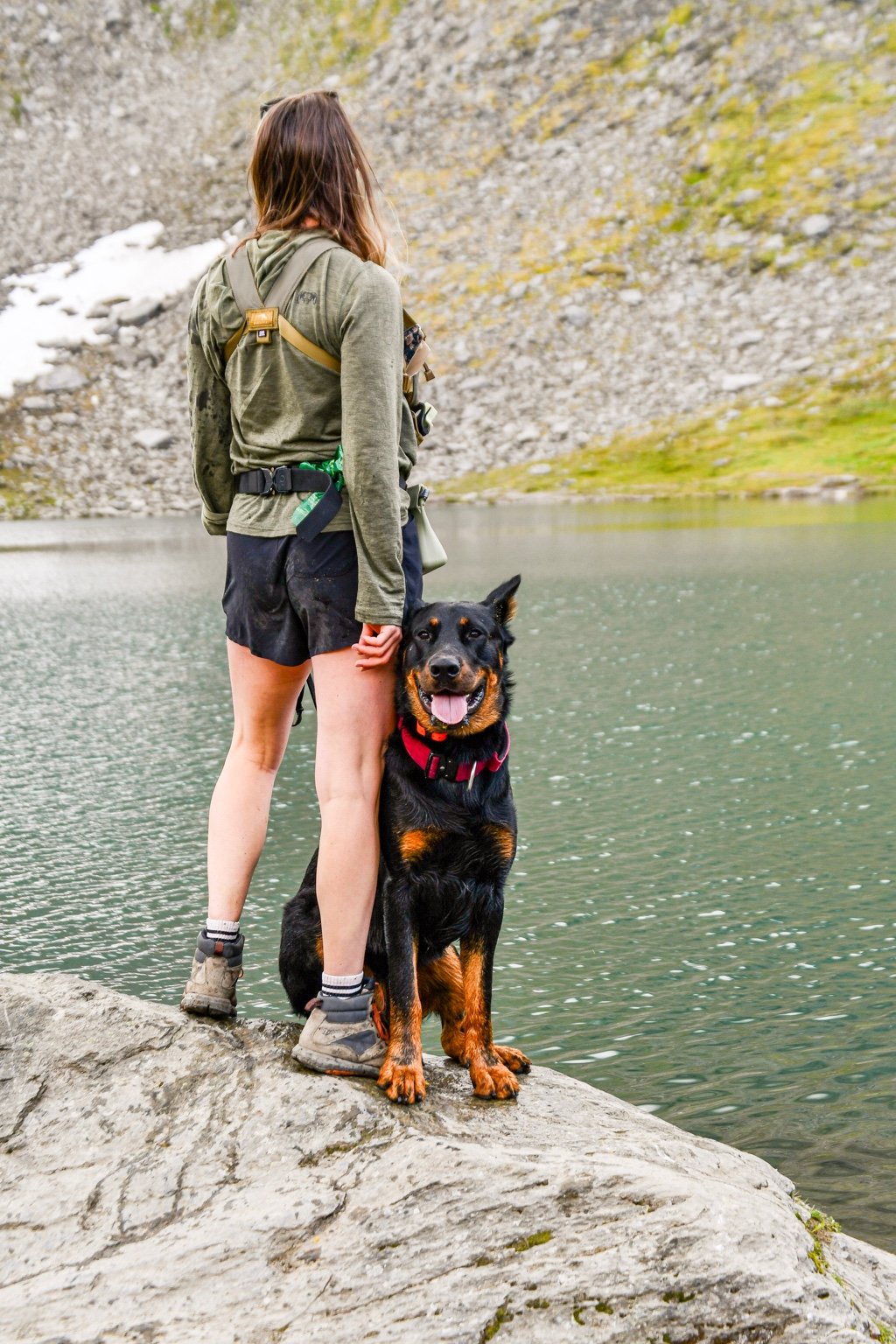 An owner and Beauceron stand together beside a mountain lake.