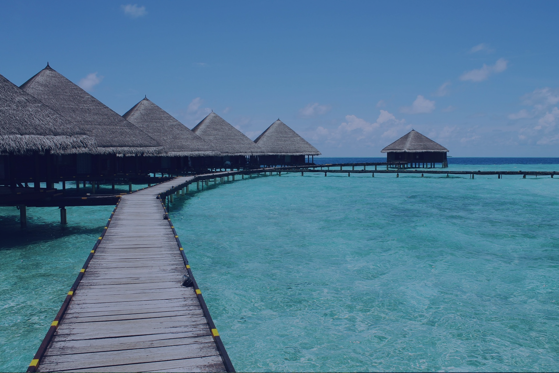 Overwater bungalows on a clear tropical ocean, connected by a wooden walkway under a blue sky are a dream. Let's start planning that dream trip for you with a Limitless Destinations luxury travel advisor.