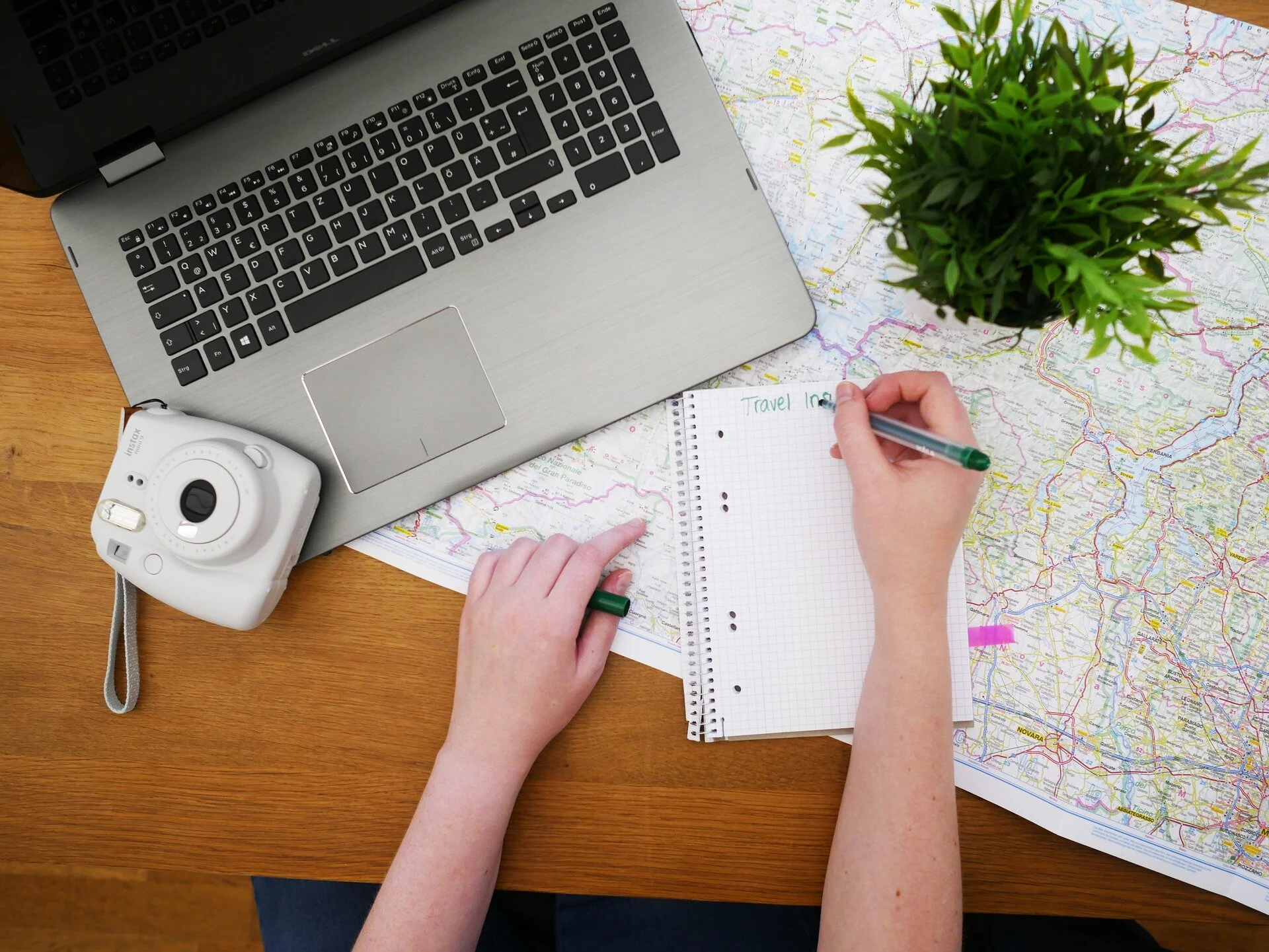 A luxury travel designer planning a bespoke journey for a client using a travel journal, a map with a laptop, instant camera, and a plant on the wooden desk.