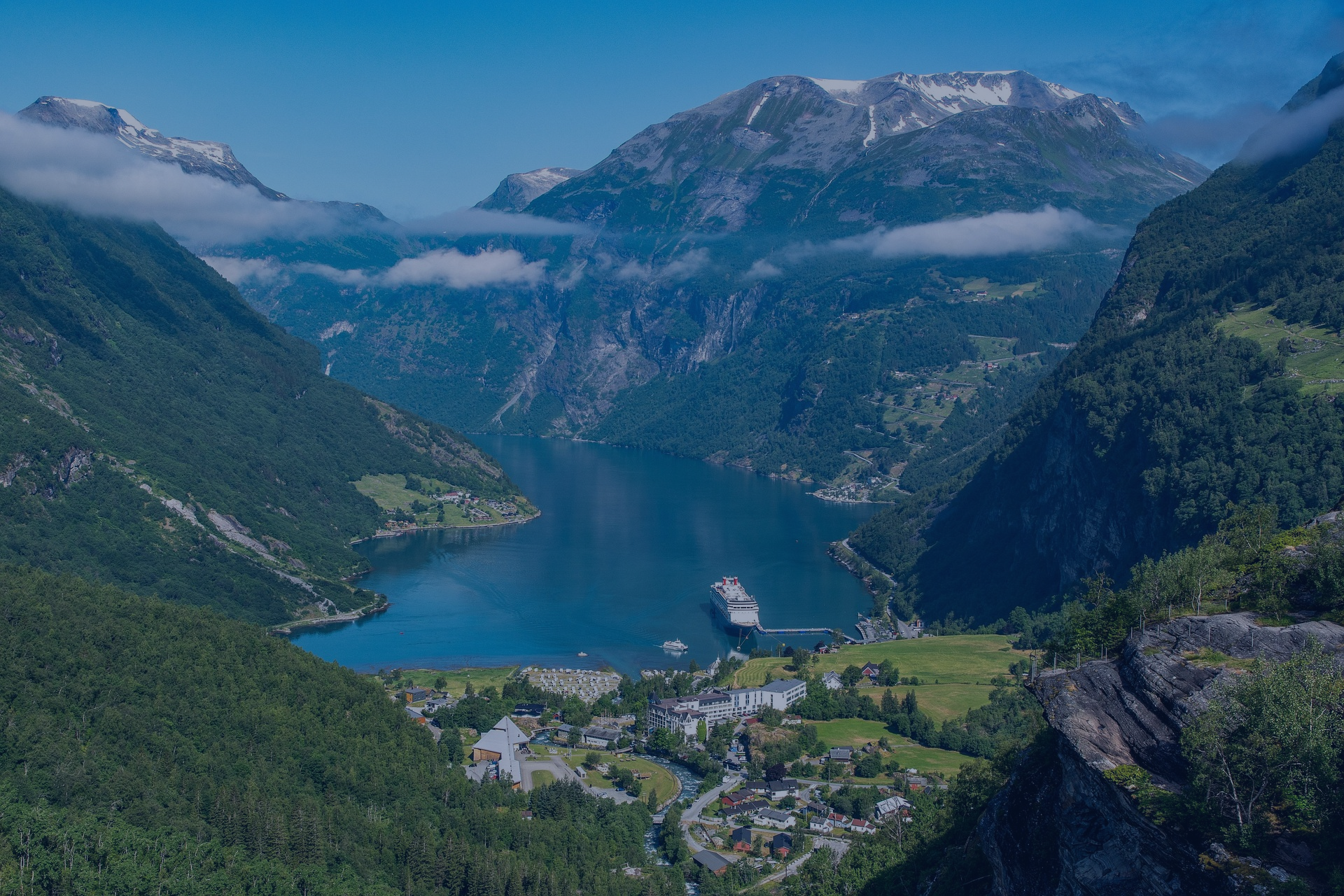 Let Limitless Destinations plan your journey to  fjord surrounded by lush green mountains with snow-capped peaks, with a cruise ship docked at the harbor, and a small village at the base of the mountains.