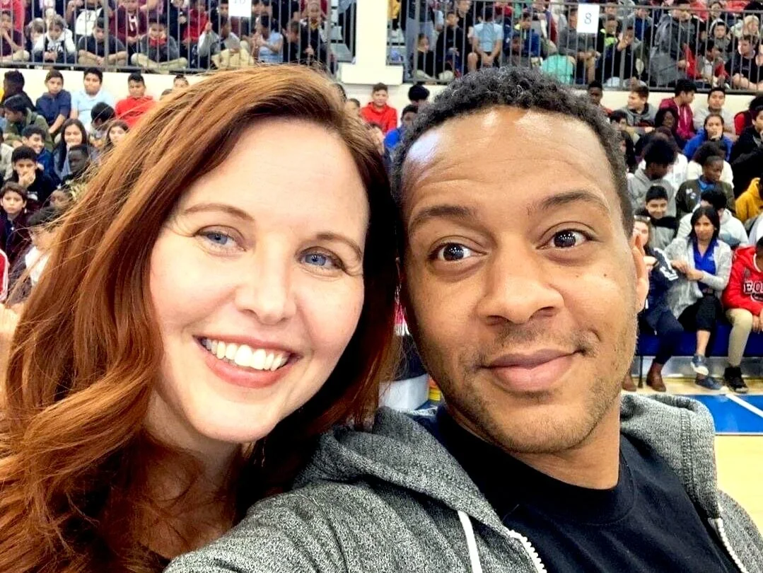 A smiling woman with red hair and a man with short curly hair taking a selfie in a crowded indoor stadium or gymnasium filled with seated spectators.
