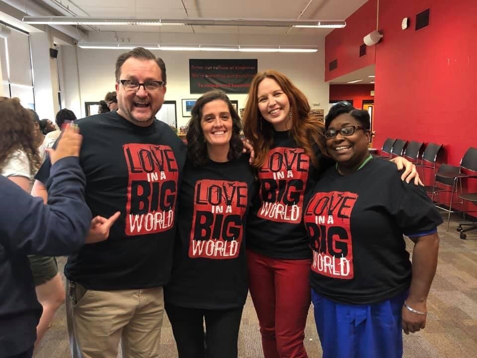 Four people standing together inside a room, smiling, wearing matching black T-shirts with red and white text that says, "Love in a Big World," with other people in the background.