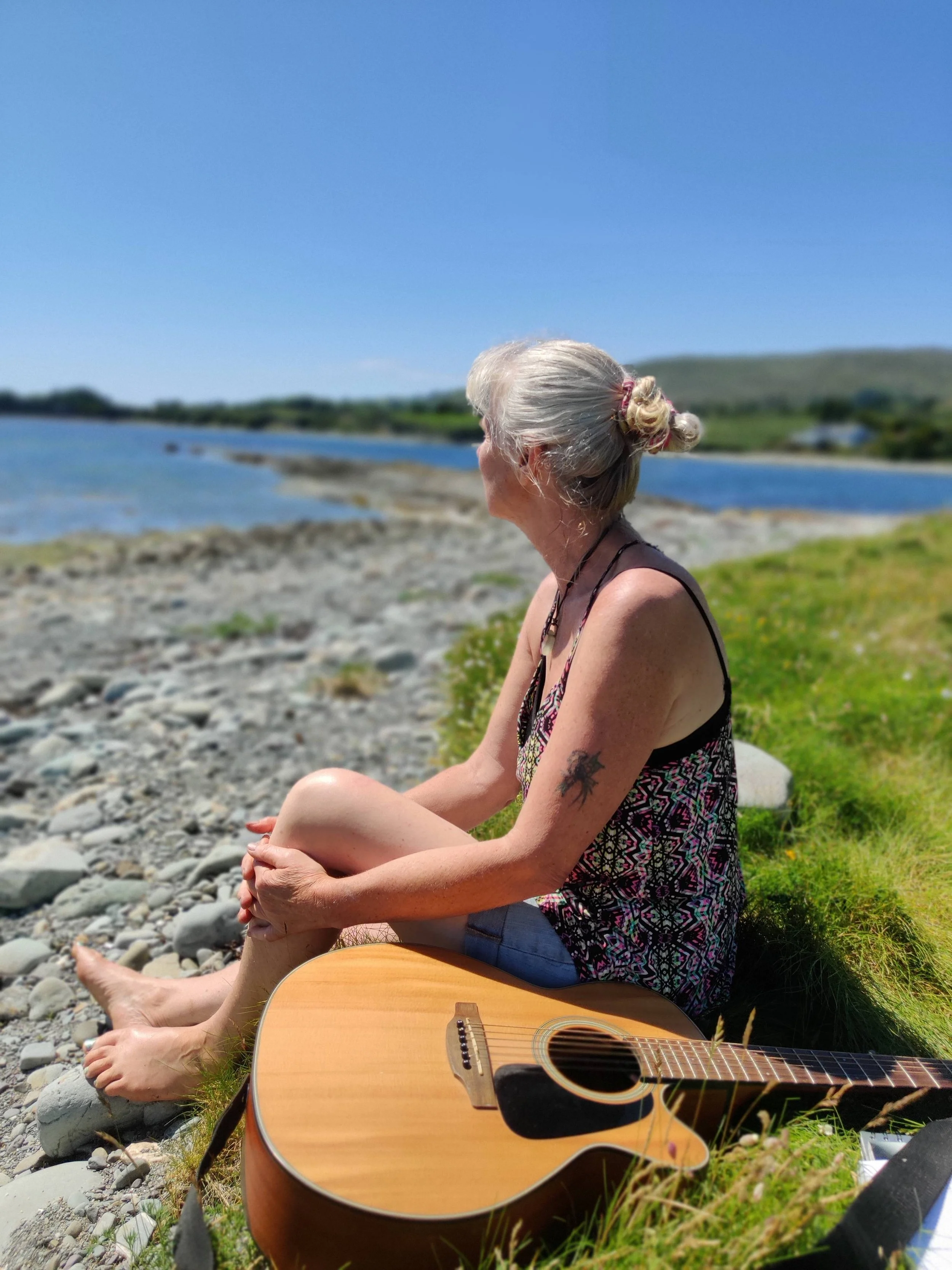 Caz is depicted by the shore seated next to her guitar while looking at the horizon