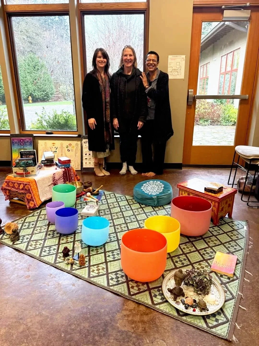 Three women standing inside a room with a large window and a glass door, smiling. In front of them is a colorful setup with large, vibrant bowls, a patterned mat, and various objects used for meditation or ceremony, including crystals, figurines, and a book.